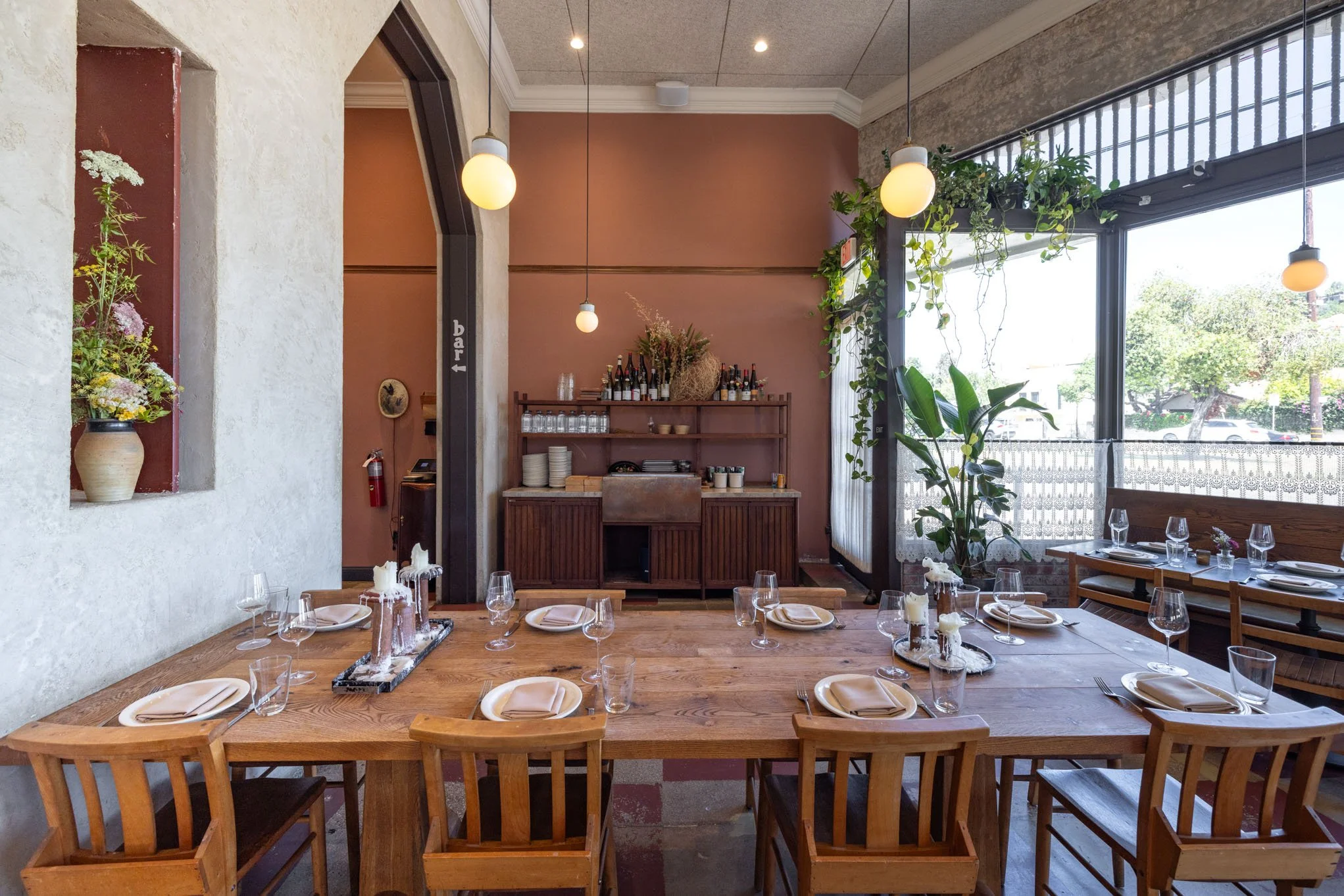 A dining setup in a restaurant with a large wooden table set with white napkins, plates, and wine glasses. There are six chairs around the table. The background features a bar area with bottles, glasses, and decor, and large windows letting in natura
