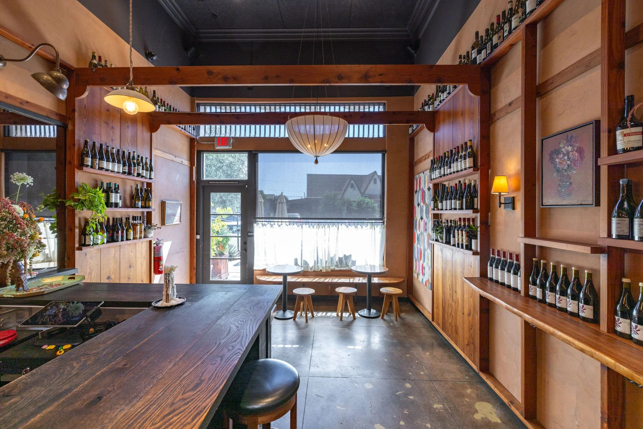 Interior of a cozy wine bar or cafe with wooden shelves filled with wine bottles, a wooden counter with a black stool, a small seating area with a table and three small stools near the window, and various decorations including a framed floral paintin
