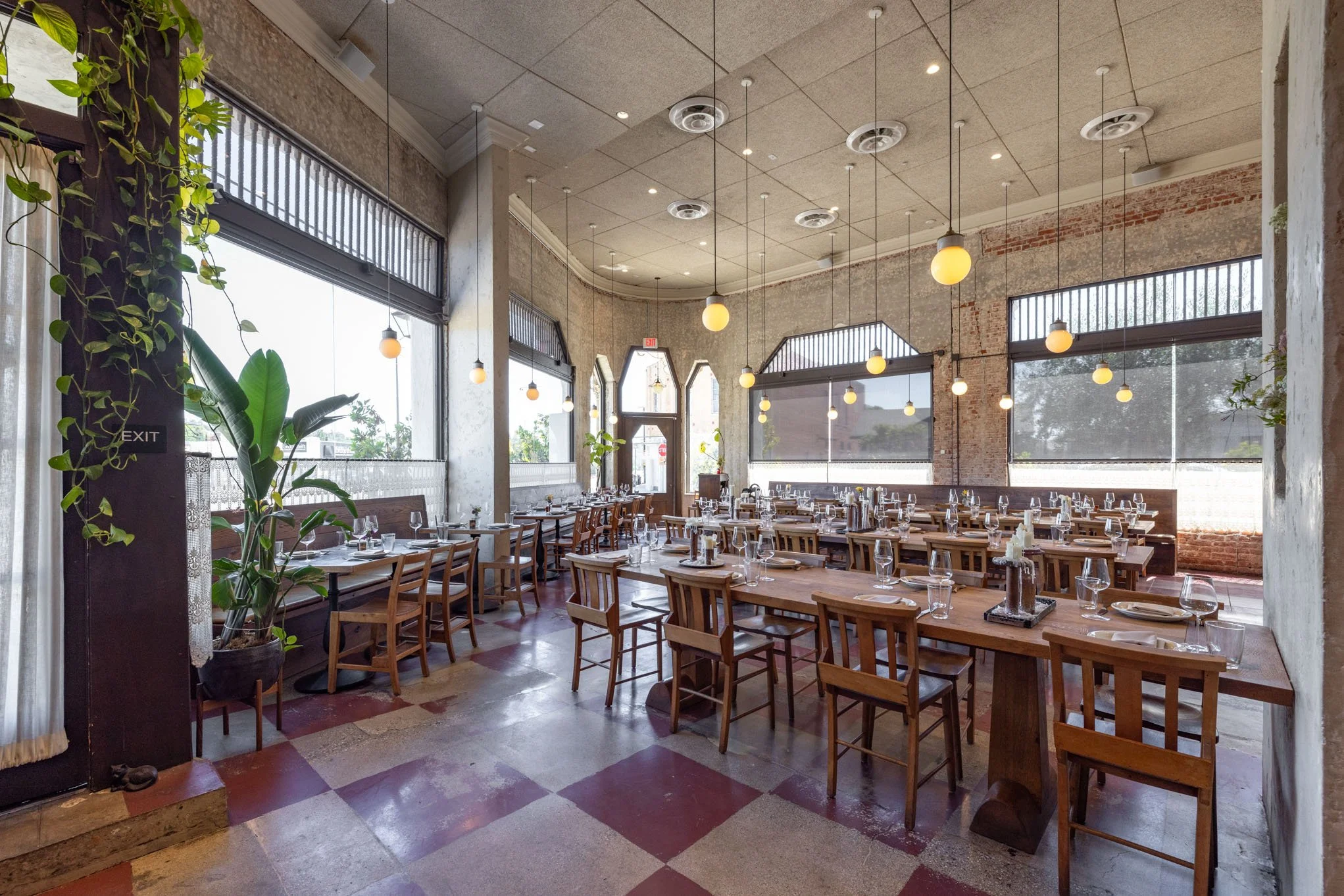 Interior of a restaurant with wooden tables set with glassware, plates, and utensils, large windows, hanging pendant lights, exposed brick and concrete walls, and potted plants.