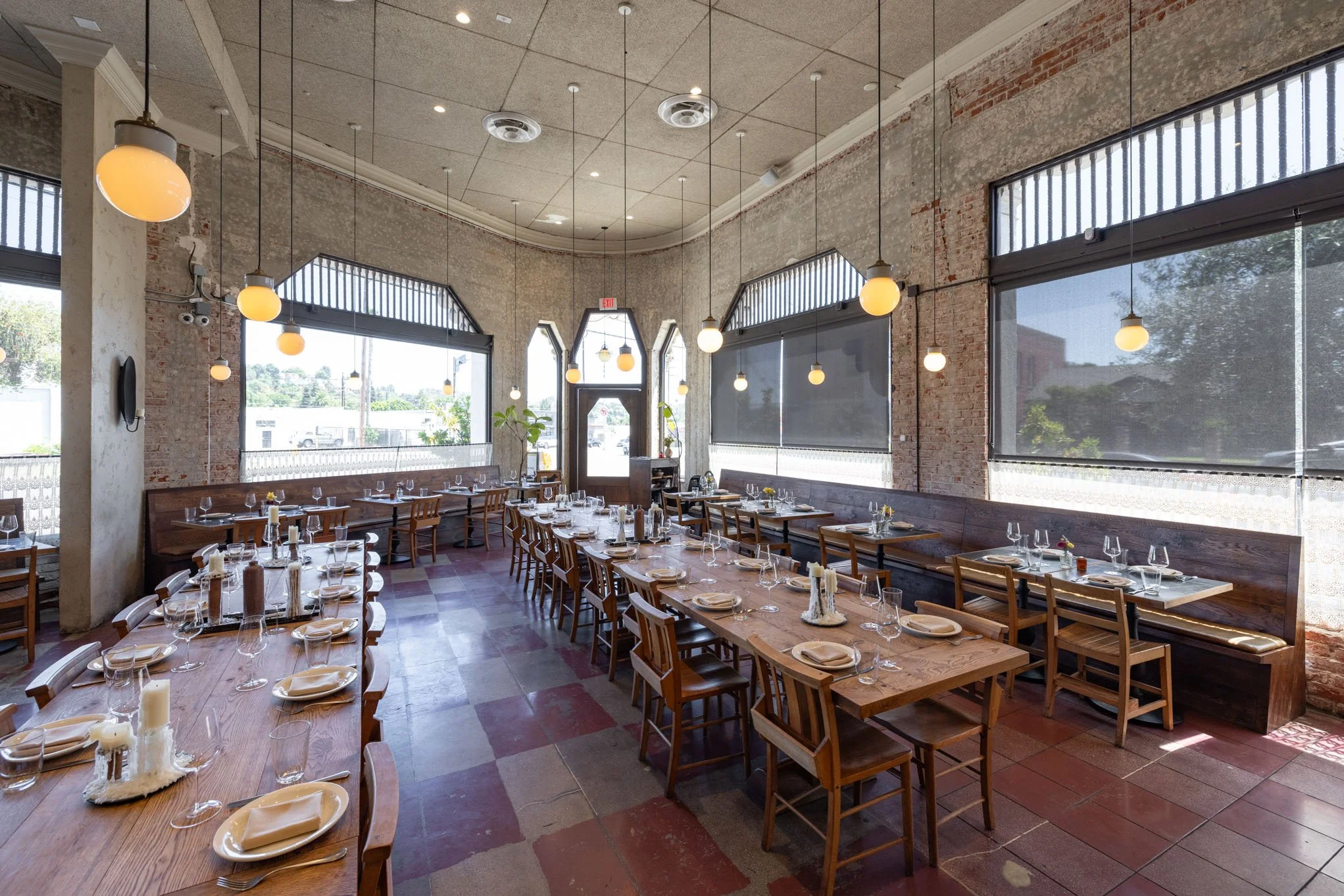 Interior of a restaurant dining area with wooden tables, chairs, and bench seating, set with plates, glasses, and candles, illuminated by hanging round light fixtures, large windows with blinds, exposed brick and concrete walls, and a tiled floor.