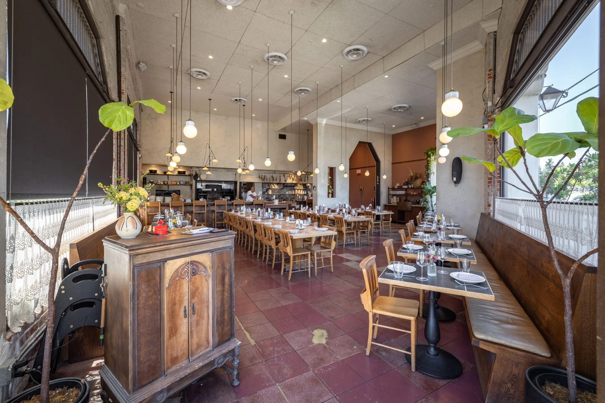 Interior of a restaurant with wooden tables set for dining, natural light from large windows, wooden chairs, decorative potted plants, and pendant lights hanging from the ceiling.