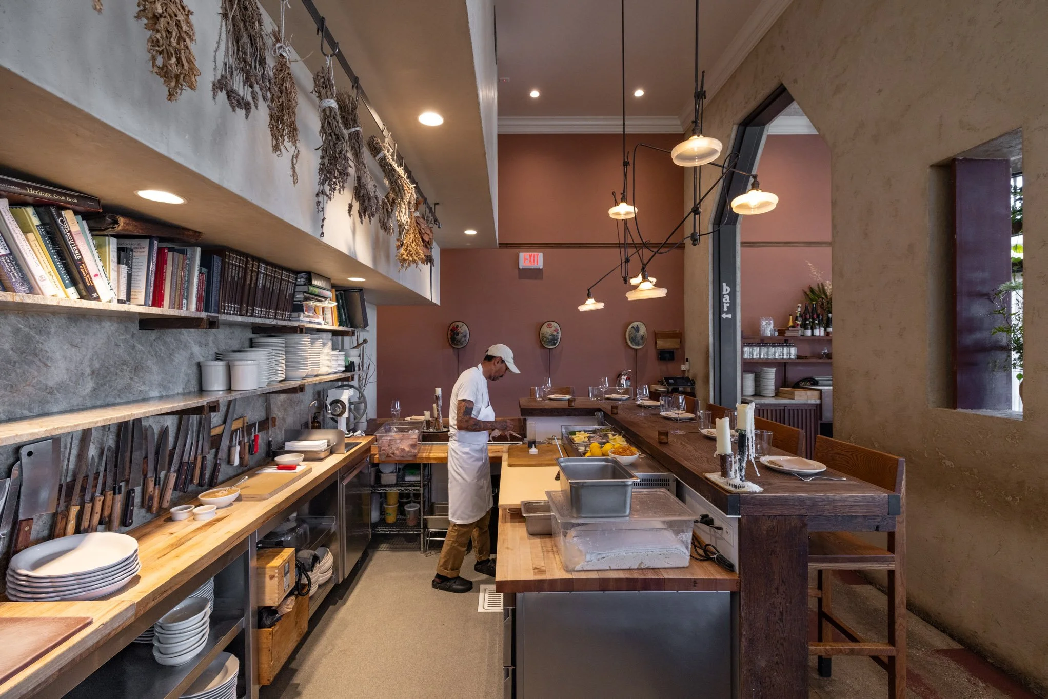 Interior of a restaurant kitchen with a chef preparing food behind a wooden counter, shelves with books, dishes, and cooking utensils, and hanging dried herbs, in a warmly lit space.