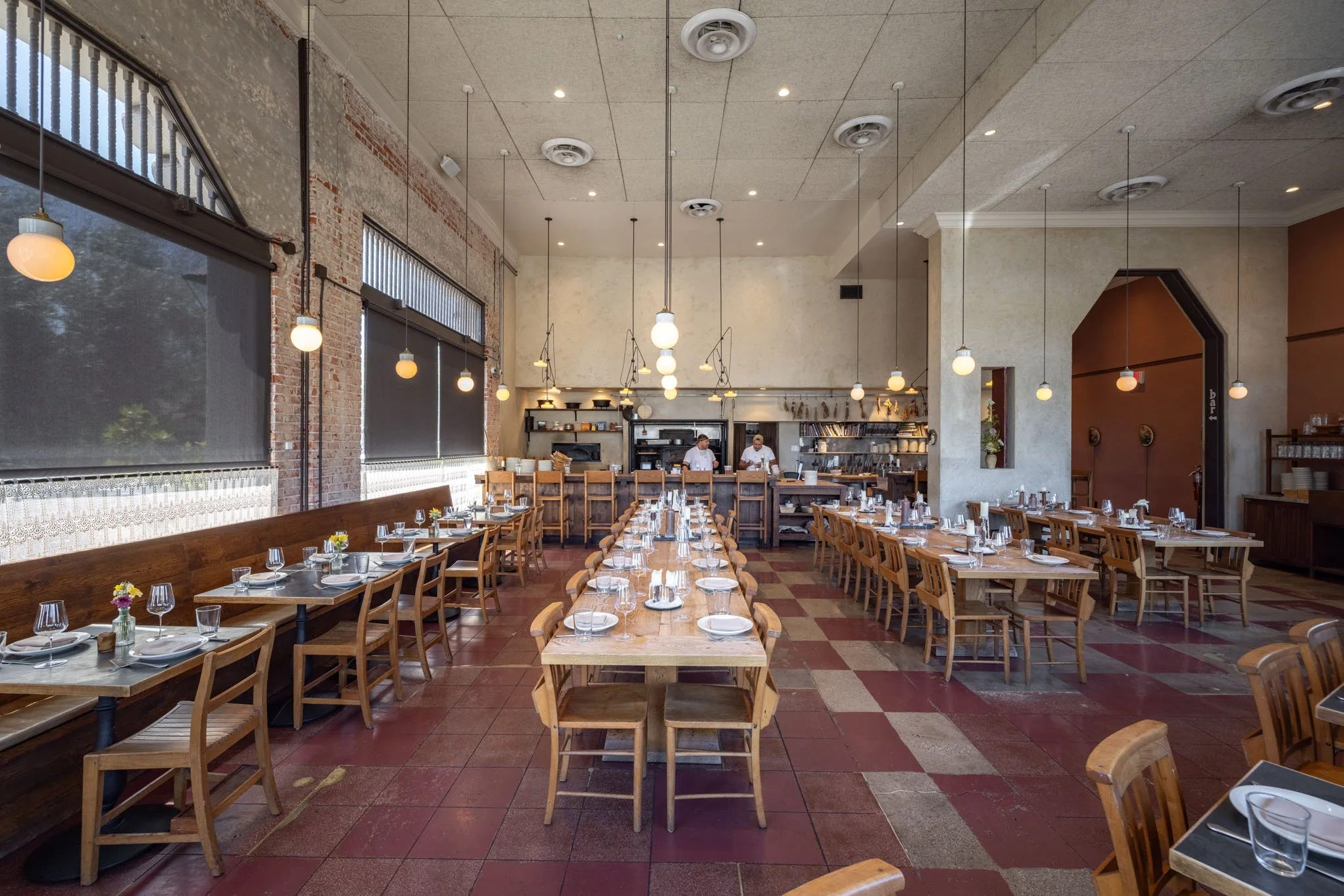 Inside a modern restaurant with wooden tables and chairs, set with glasses, plates, and silverware. The space has large windows with black blinds, exposed brick and a beige wall, pendant lights hanging from the ceiling, and a kitchen in the backgroun