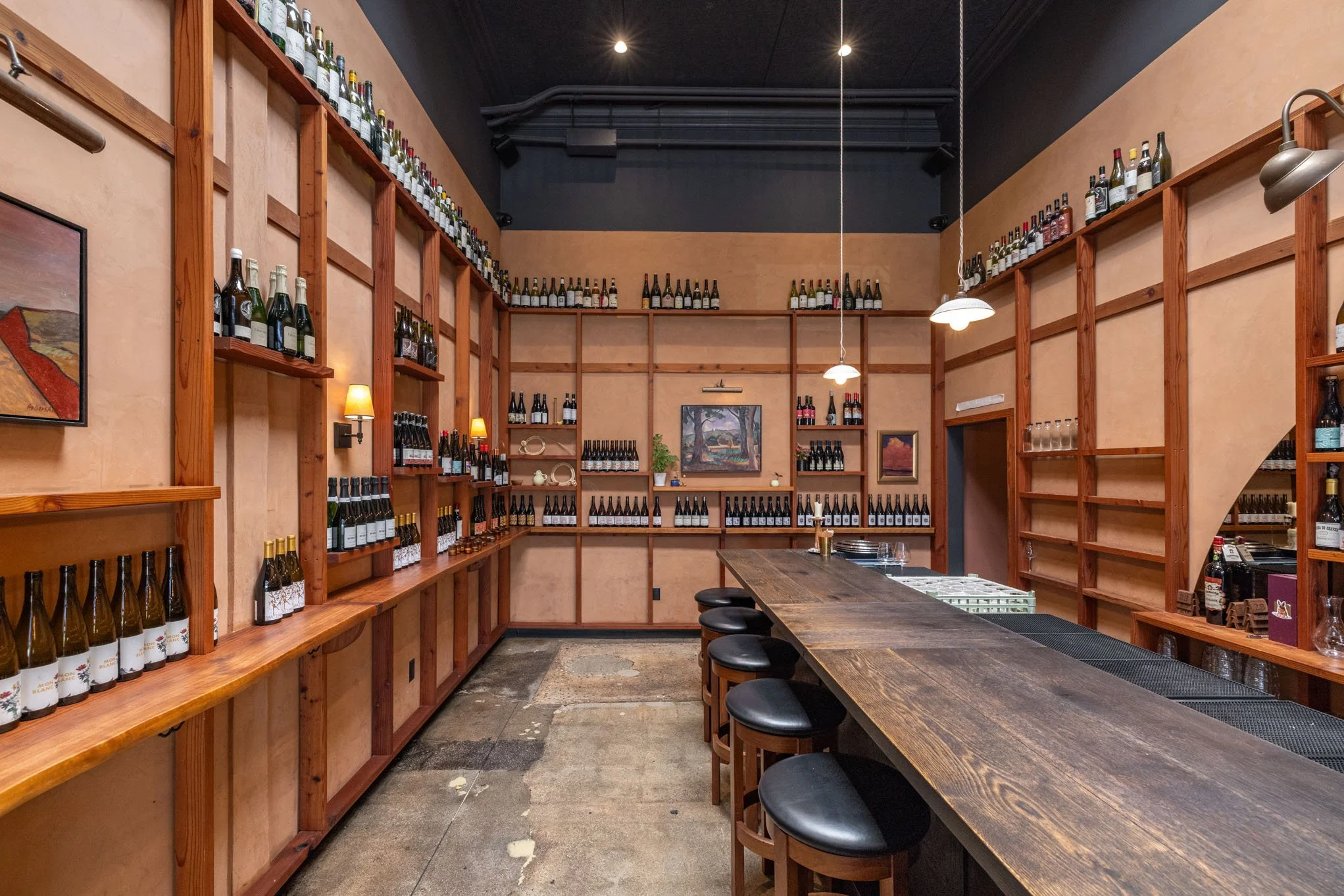 Interior of a wine bar with shelves of wine bottles along the walls, a long wooden bar counter with bar stools, and framed artwork on the walls.