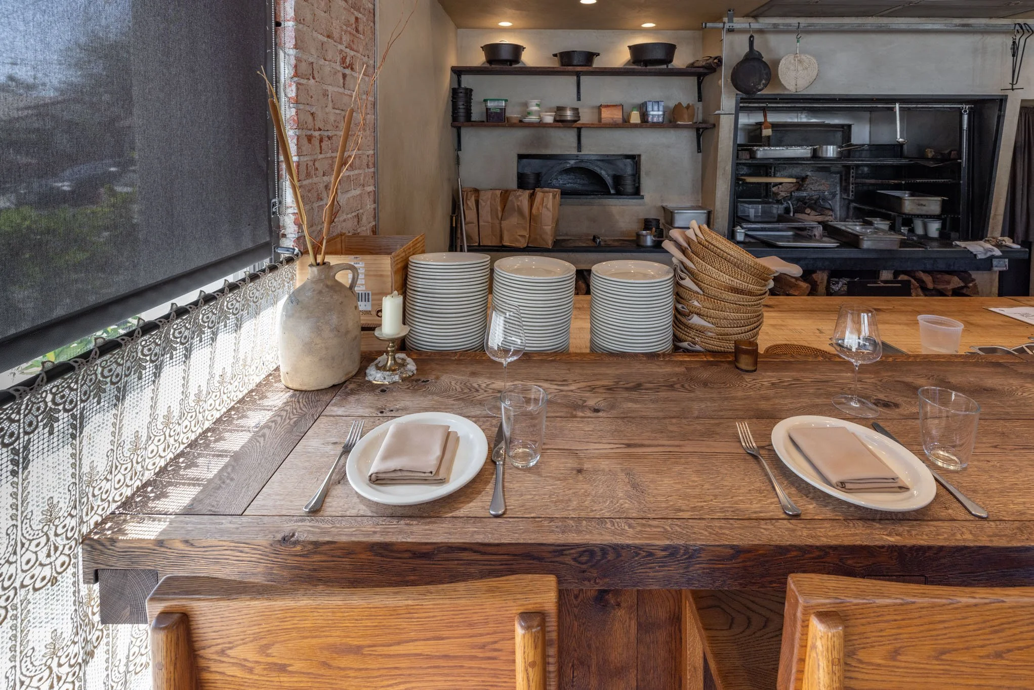 A rustic restaurant interior with a wooden dining table set with napkins, glasses, and utensils, and an open kitchen in the background.