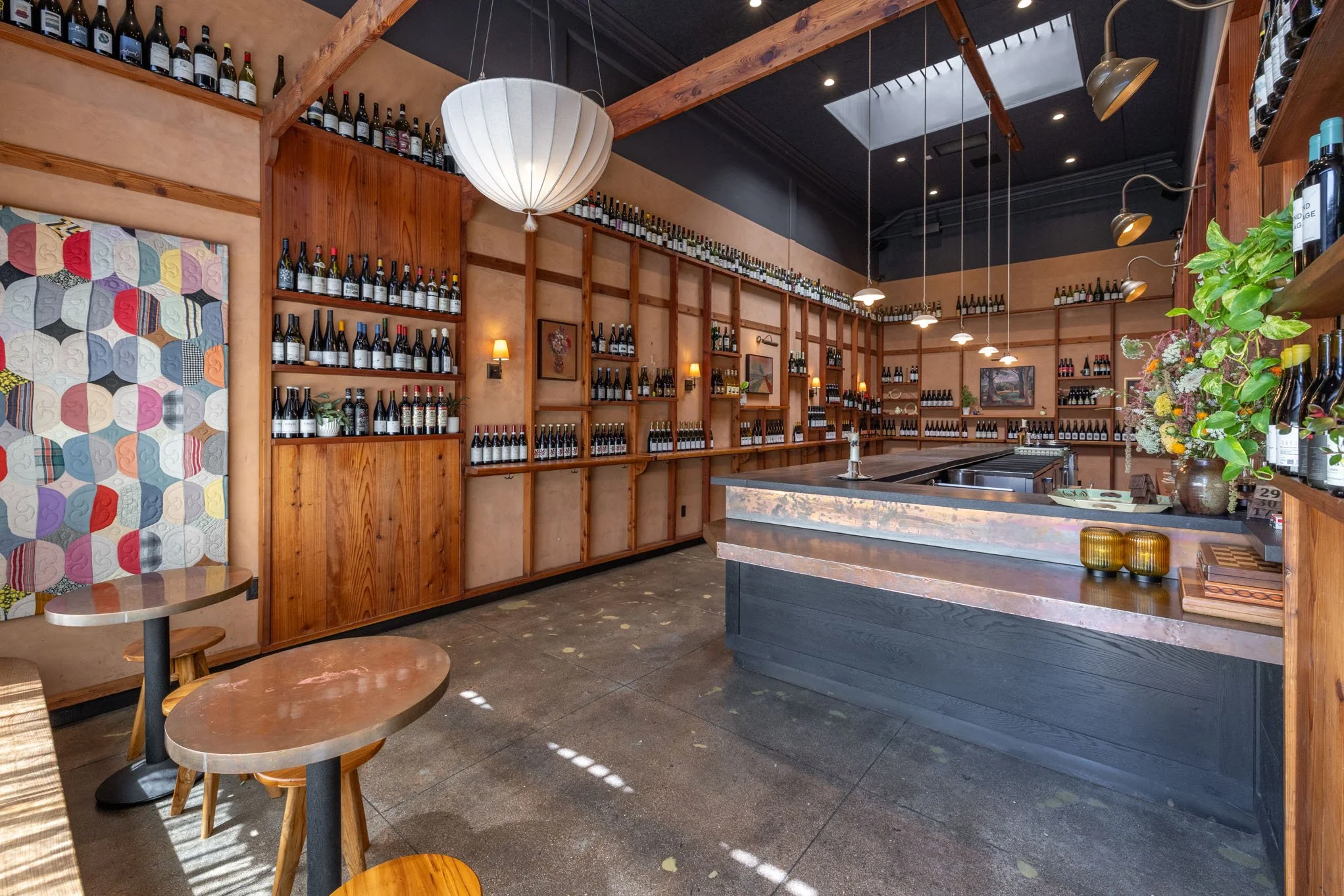 Interior of a modern wine bar with numerous bottles of wine on wooden shelves, a colorful quilt hanging on the wall, and a counter with a vase of flowers and candles.