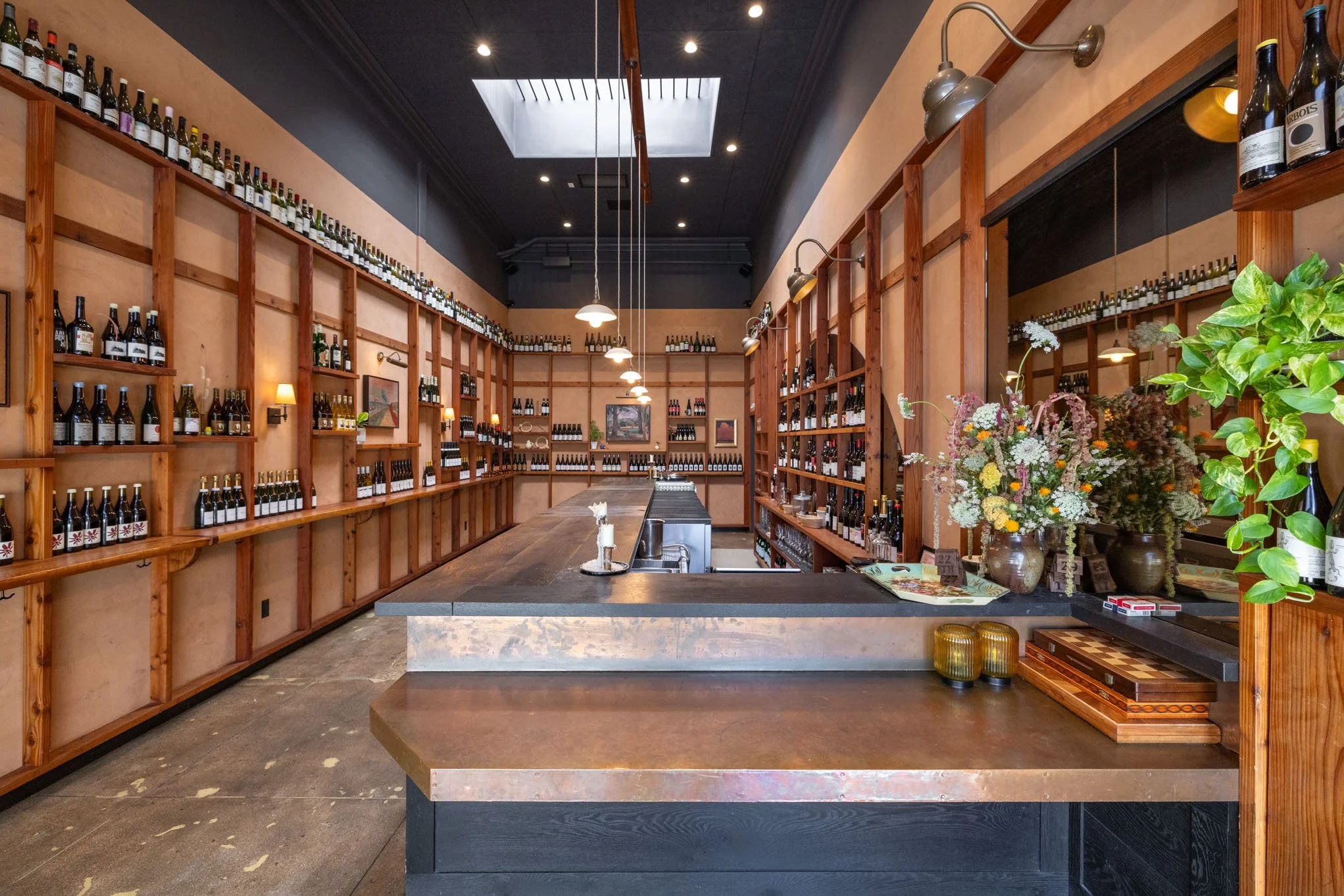 Interior of a wine shop with wooden shelves filled with wine bottles, a long wooden counter with a flower arrangement, and small light fixtures on the walls.