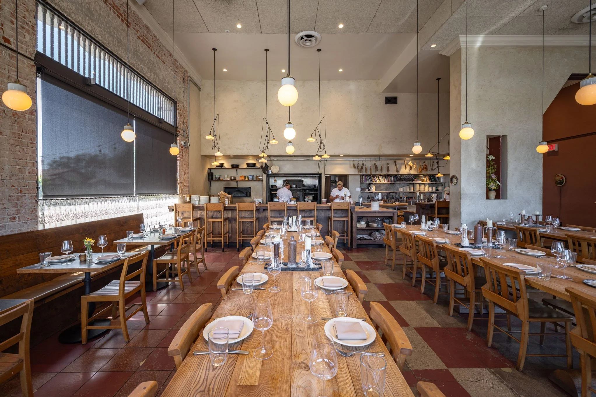 A cozy restaurant interior with wooden tables and chairs, set for dining with place settings, glasses, and small flower arrangements. The open kitchen at the back has chefs preparing food, and large windows with blinds provide natural light. The atmo