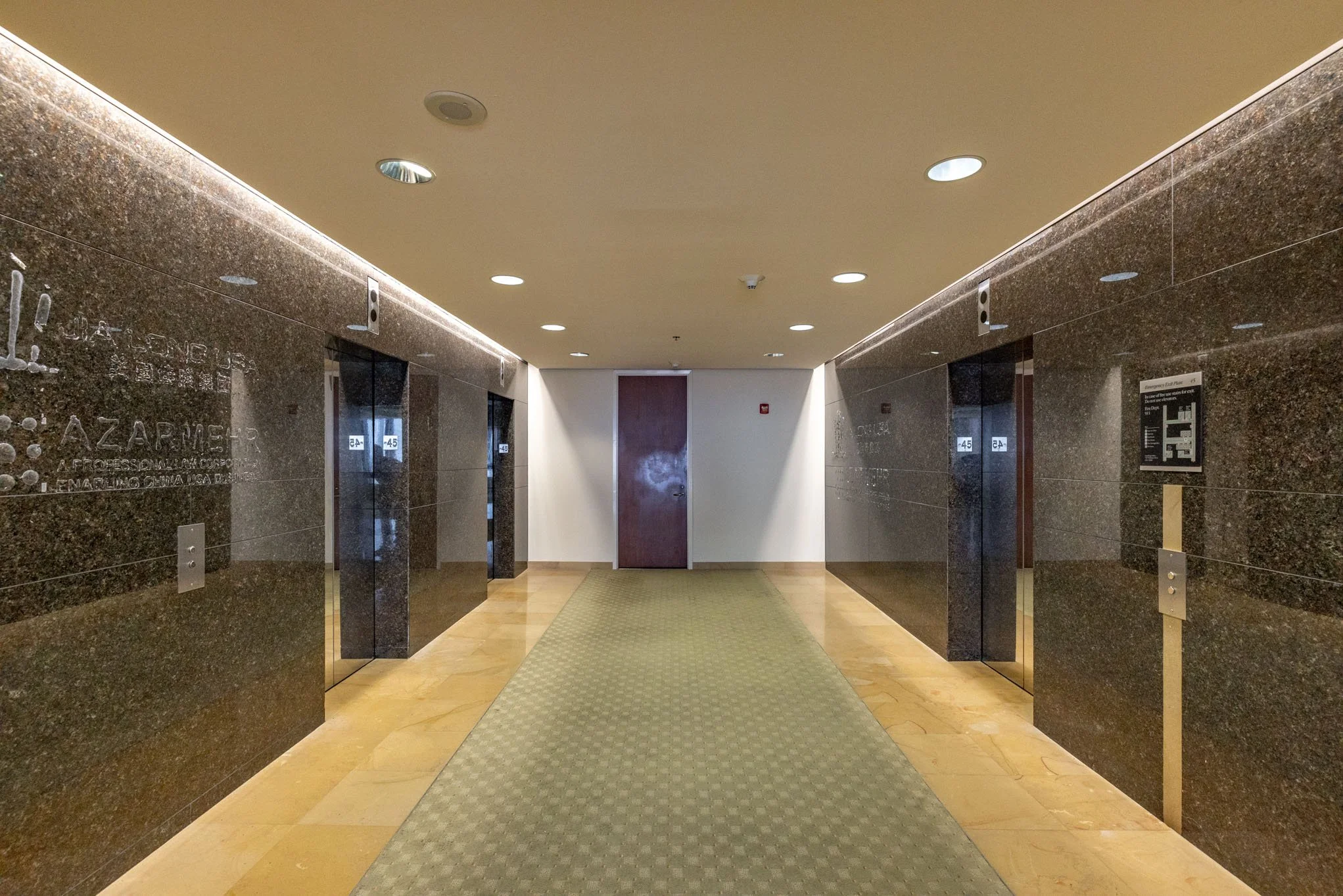 Empty elevator lobby with marble walls, two elevators, a green carpeted floor, and a closed wooden door at the end.
