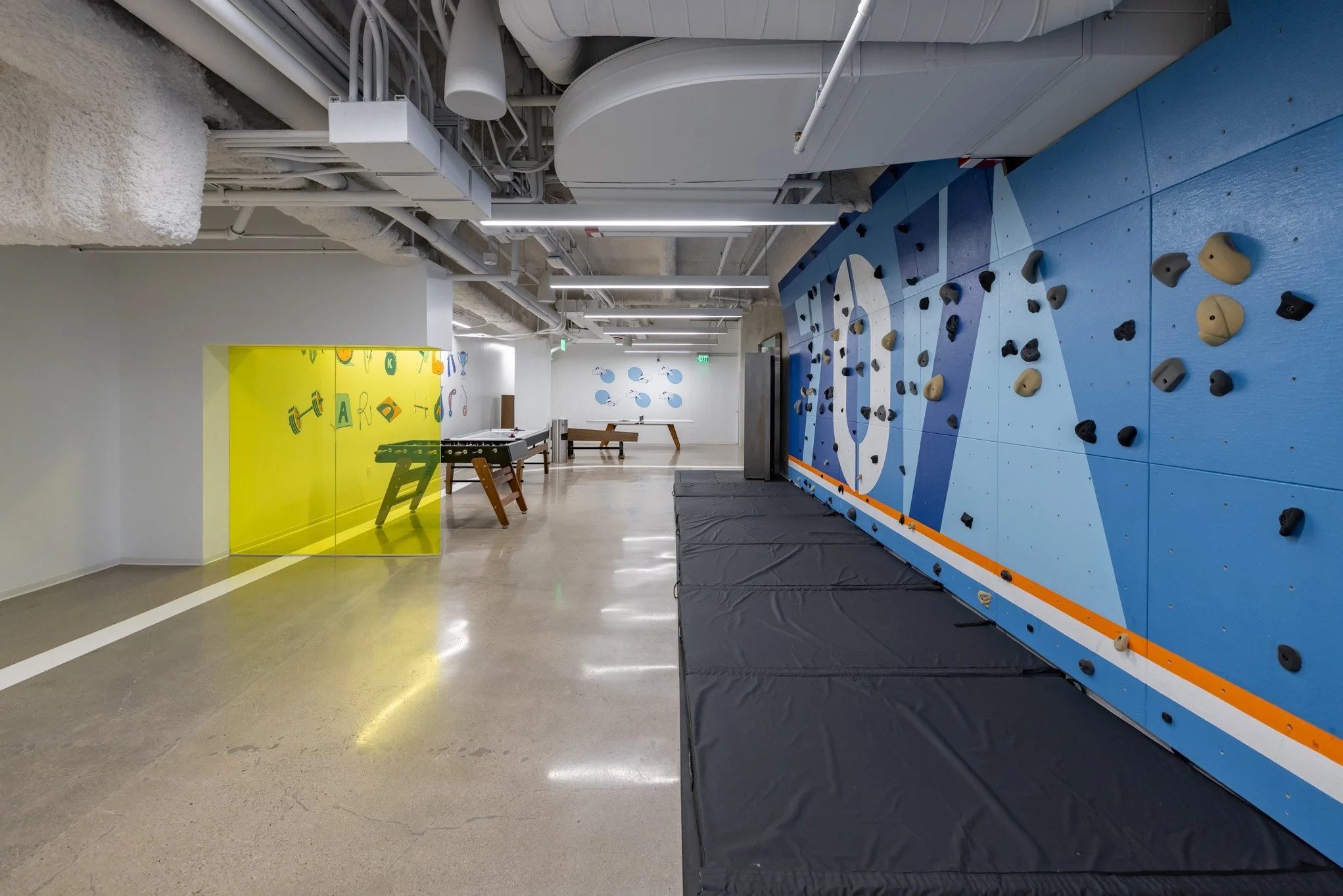 Indoor rock climbing gym with a blue climbing wall on the right and a yellow wall with graphics of weights on the left, both walls equipped with climbing holds. The floor is shiny with a safety mat underneath the climbing wall, and there are benches 