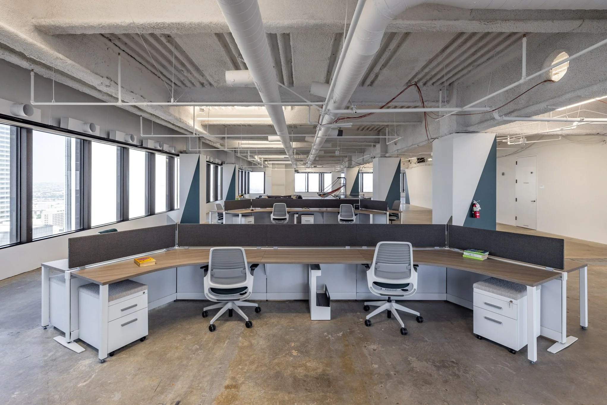 Modern empty office with large windows, gray desks with dividers, office chairs, and exposed ceiling with ductwork and pipes.