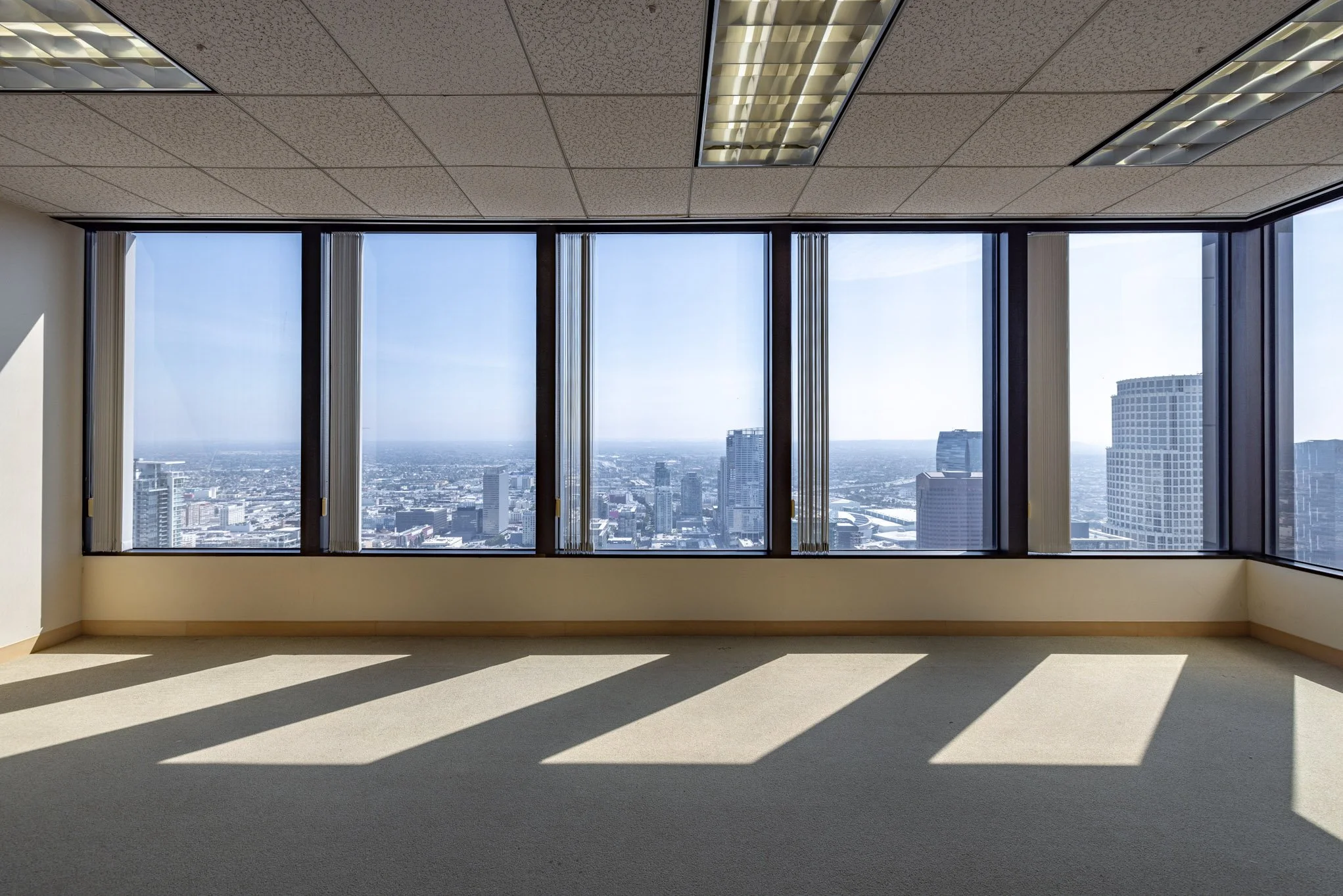 Empty office room with large floor-to-ceiling windows showing a city skyline, sunlight casting shadows on the beige carpeted floor, and a tiled ceiling with fluorescent lights.