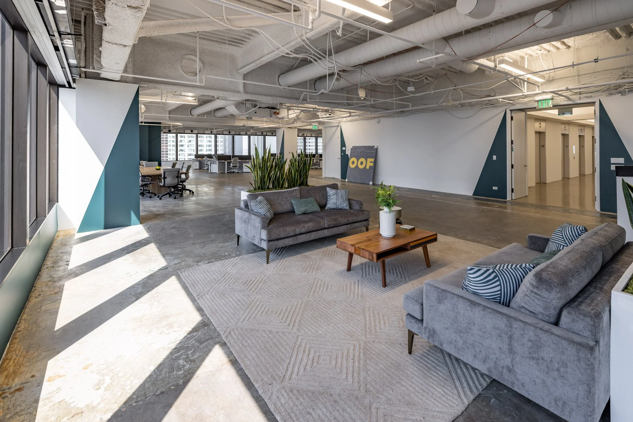 Modern office lounge area with two gray sofas, a wooden coffee table, green plants, and a beige area rug, with workstations and elevator visible in the background.
