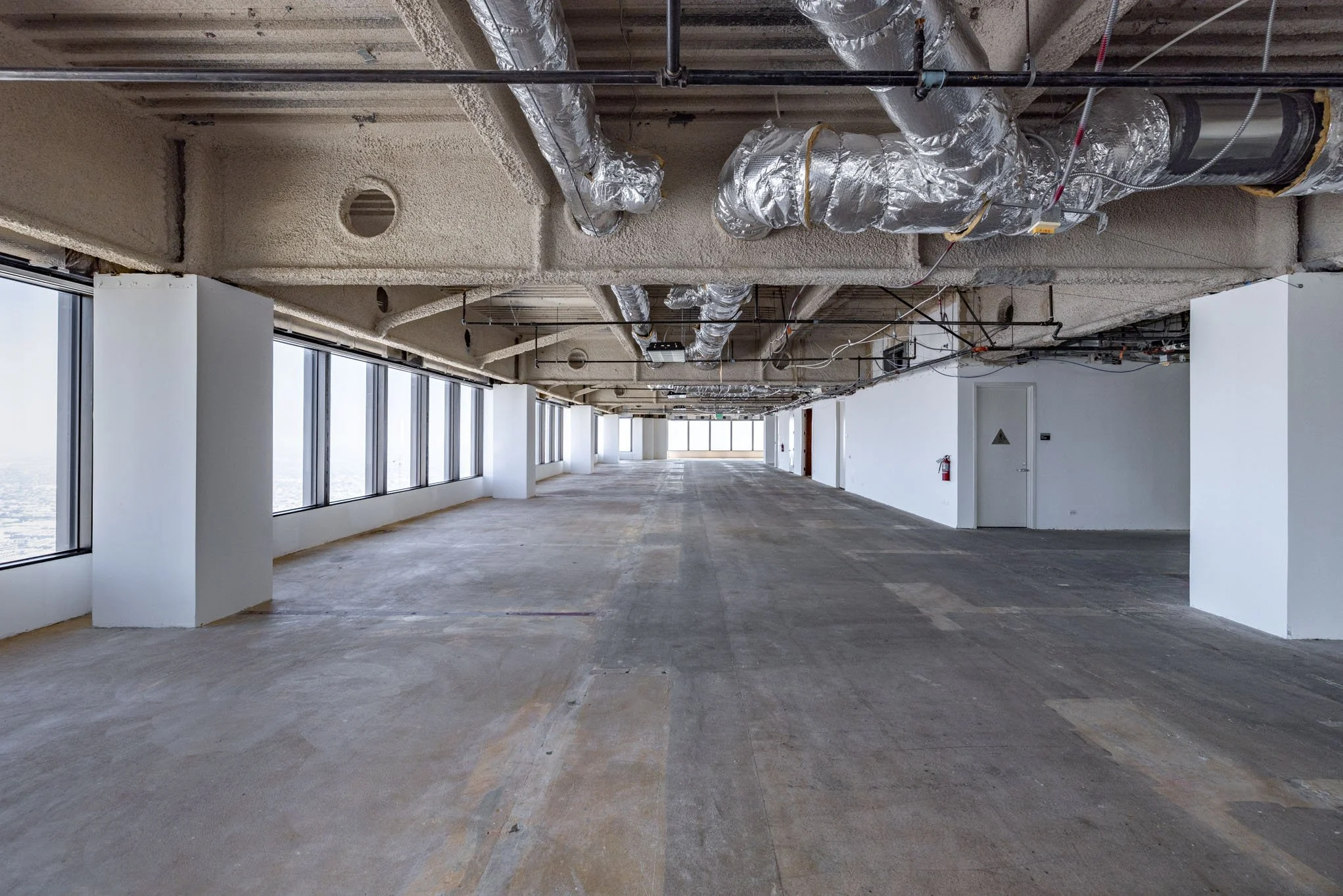 Empty office space under construction with large windows, exposed ductwork, and a fire extinguisher on the wall.