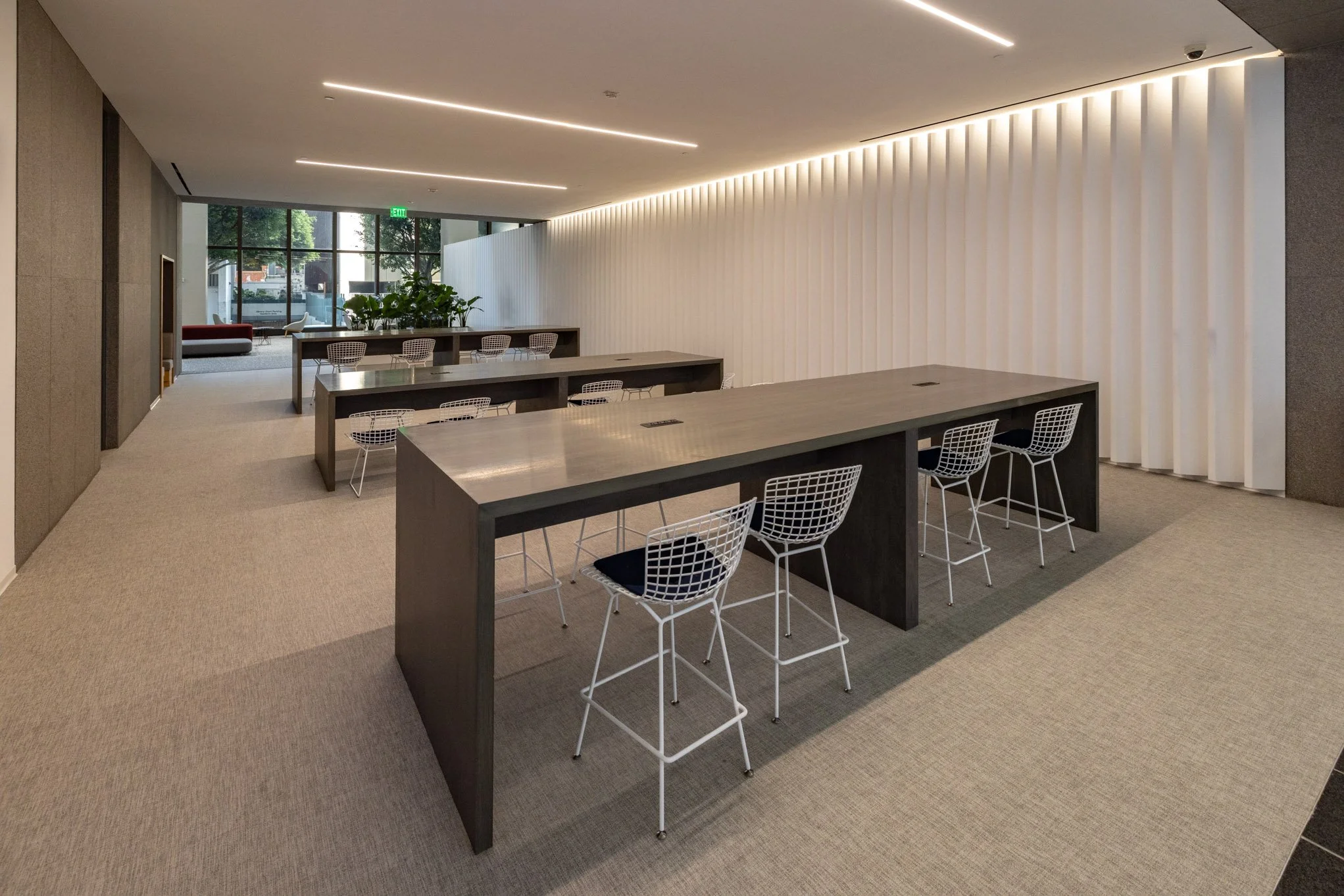 Modern interior room with long dark wood tables, white wire chairs with black cushions, beige carpet, white ceiling with recessed lighting, large window with greenery outside, and a white textured wall with vertical slats.