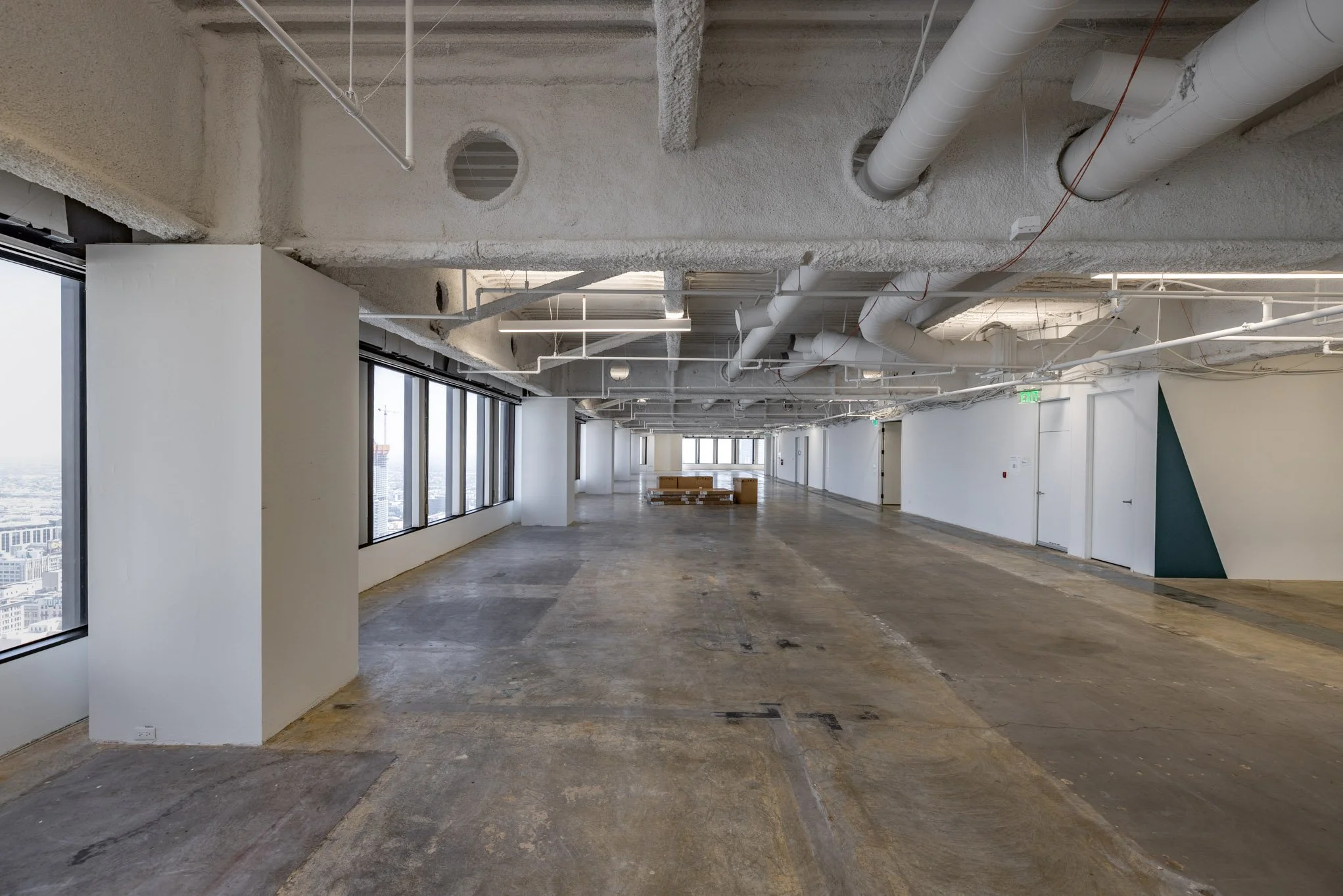 Empty office space under renovation with large windows, exposed ceiling pipes, and unfinished flooring.
