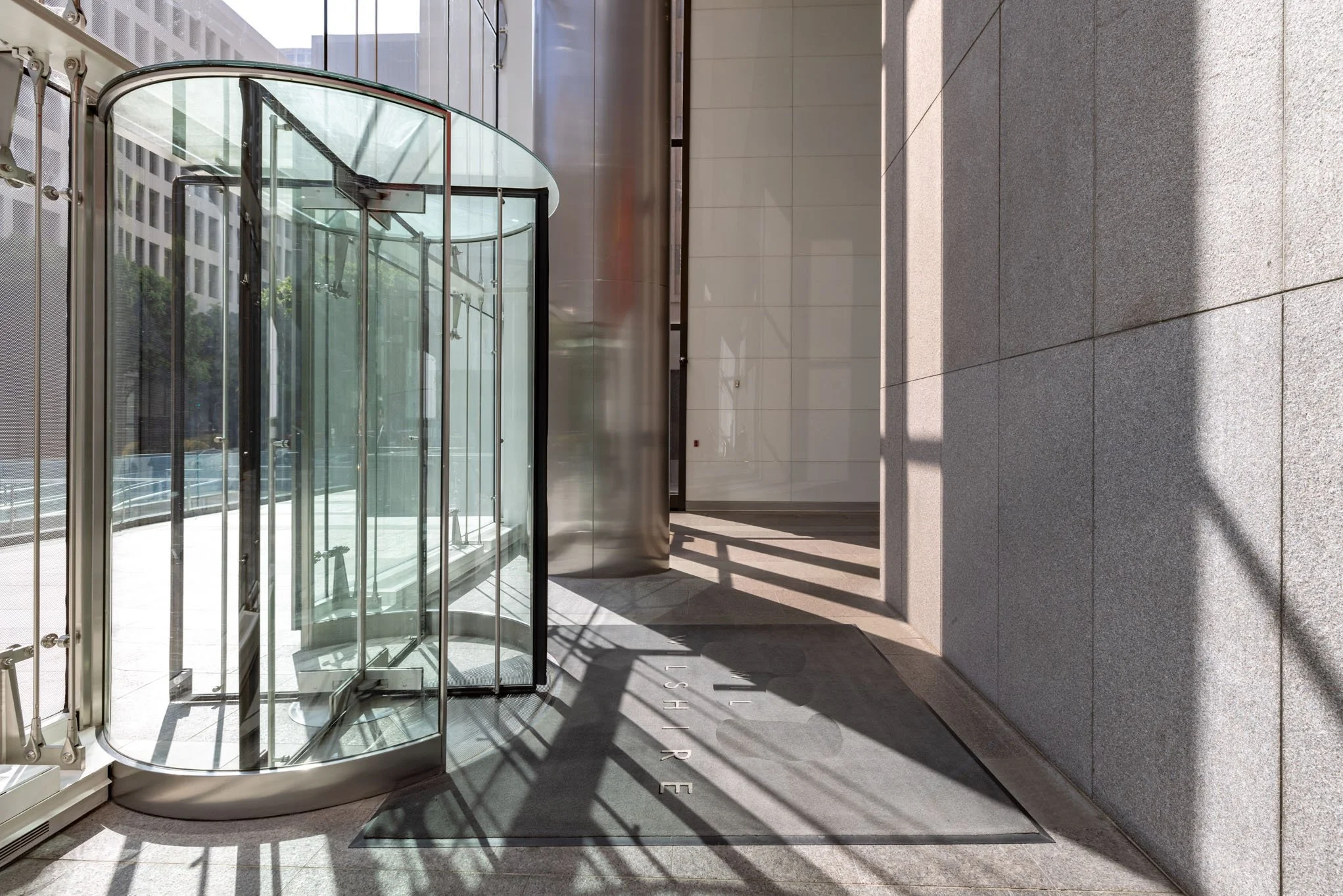 Modern glass revolving door on a city building entrance, with glass panels, metal framing, and a gray tile sidewalk in sunlight.