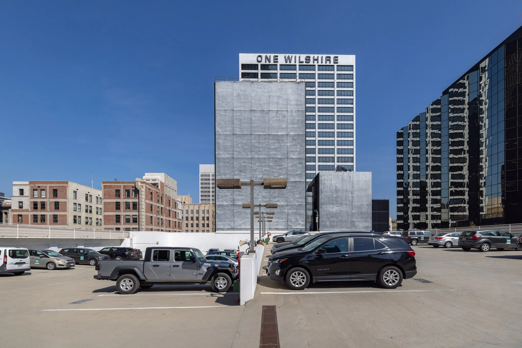 A parking lot on the rooftop of a building with cars parked. In the background, there are tall buildings, including one with a sign that reads 'One Wilshire'.