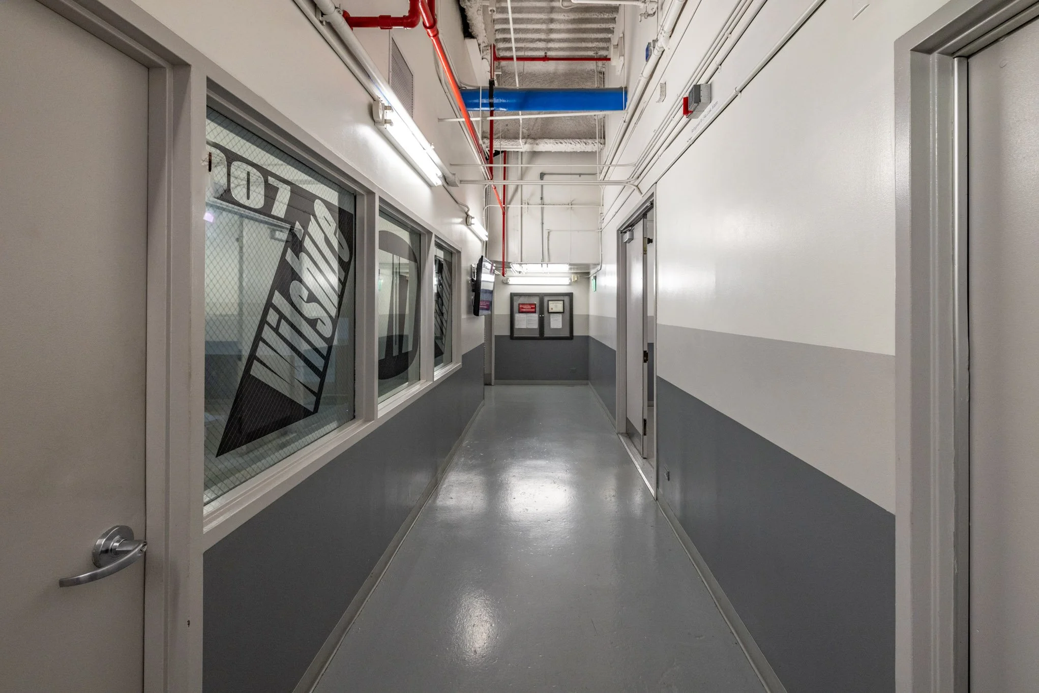 Empty industrial hallway with gray and white walls, fluorescent lights, pipes on the ceiling, and a window with the word  "VISITORS" printed on it.