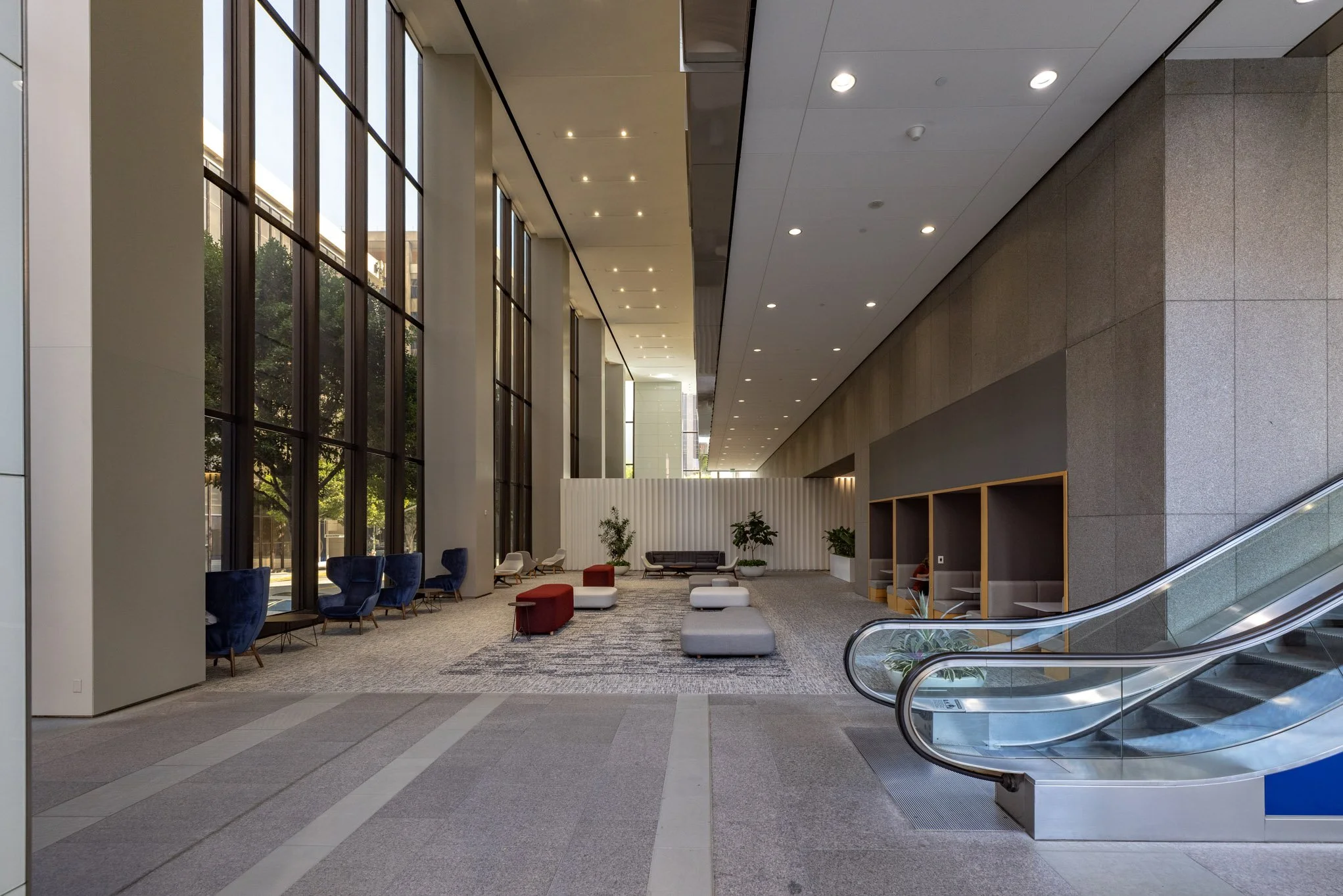 Spacious modern lobby with large floor-to-ceiling windows, seating area with blue and gray chairs, ottomans, and green plants, illuminated by ceiling lights.