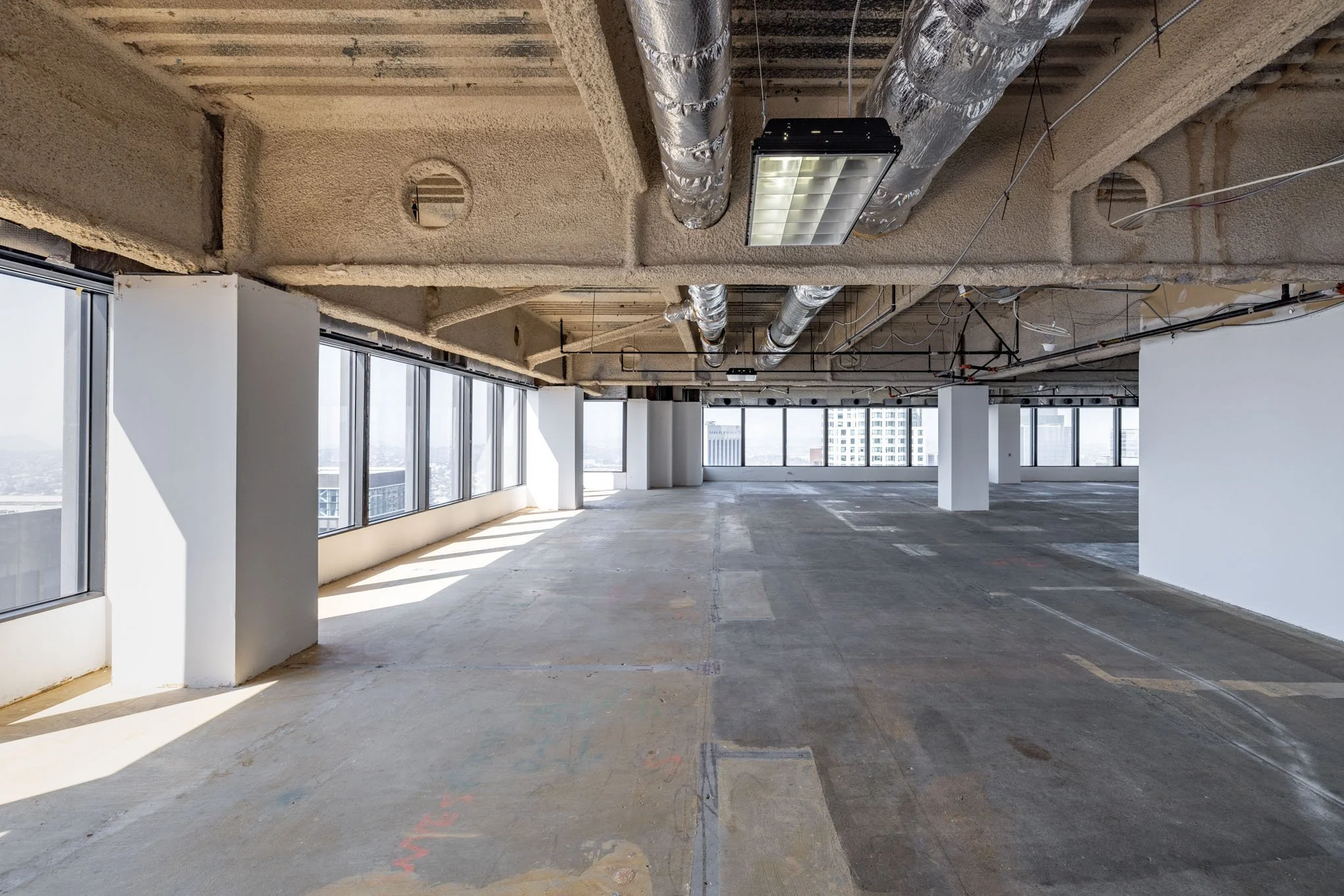 Empty commercial office space under construction with large windows, exposed ceiling with ducts and lighting, and concrete flooring.