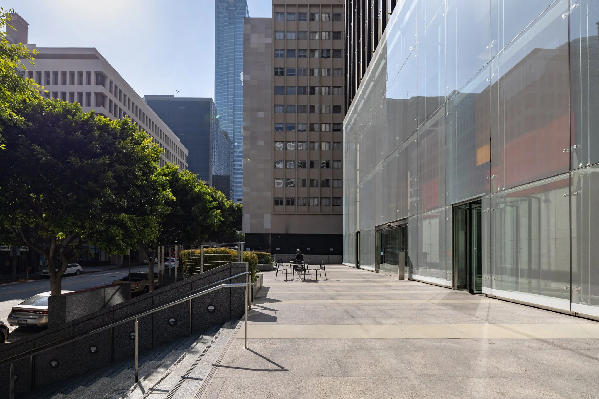 Urban street scene with modern glass building on the right, trees and parked cars on the left, and tall office buildings in the background.