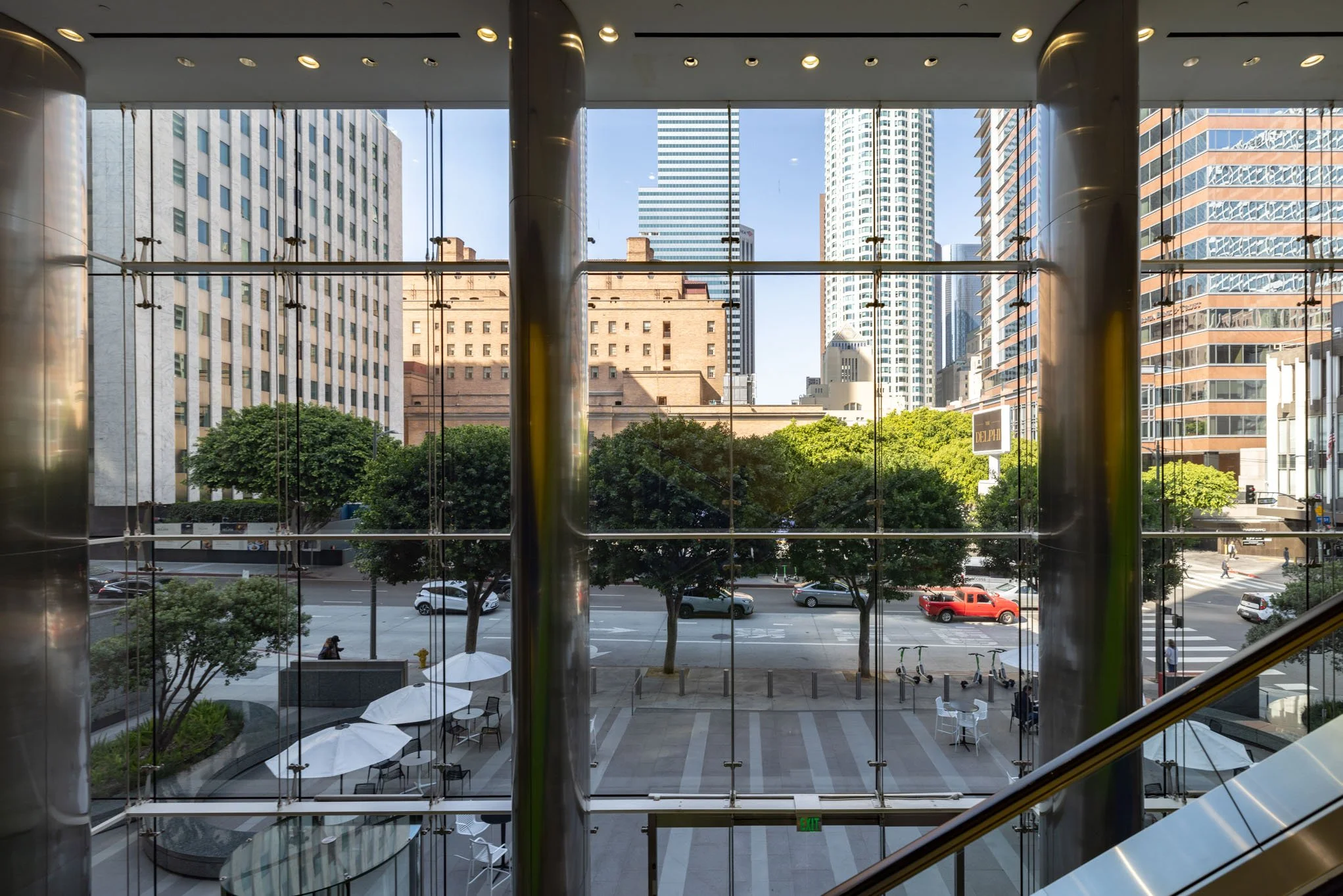 View of a city street through large glass windows from inside a building, with buildings, trees, and cars visible outside, and a terrace with umbrellas and tables inside.