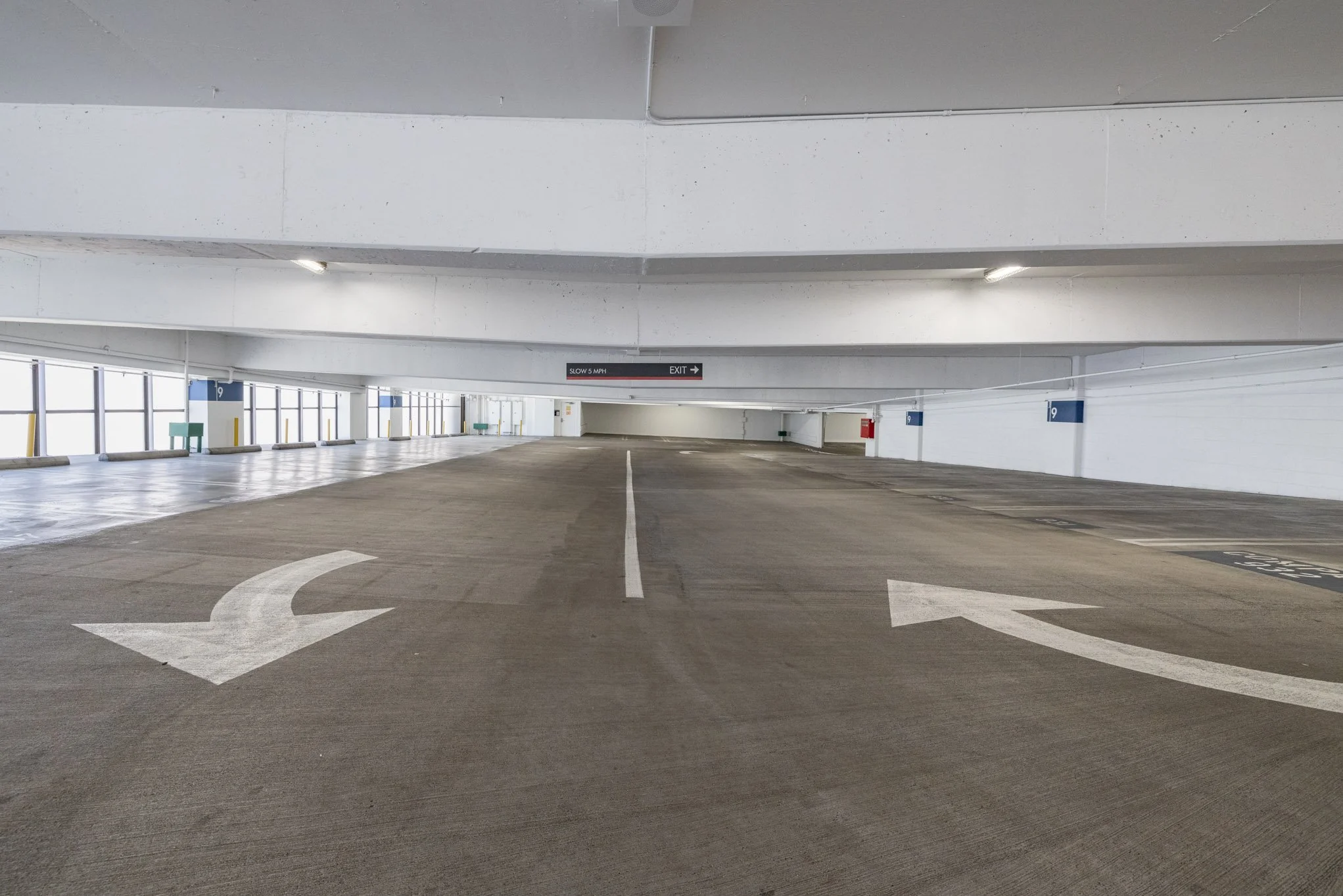 Empty multi-level parking garage with directional arrows on the pavement, concrete walls, and signs indicating parking directions and speed limit.