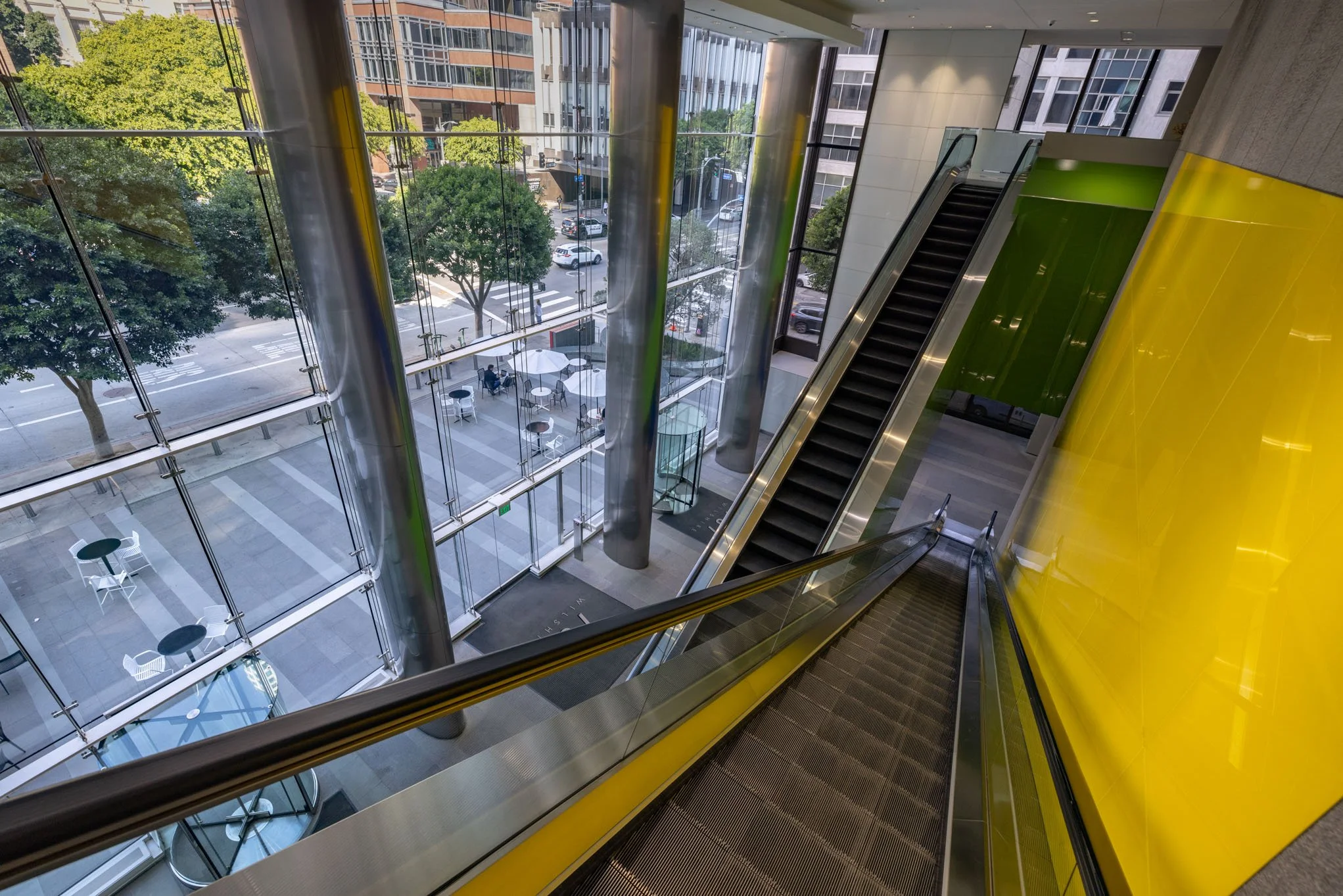 View from above of an escalator inside a modern building with large glass windows showing a city street with trees and parked cars.