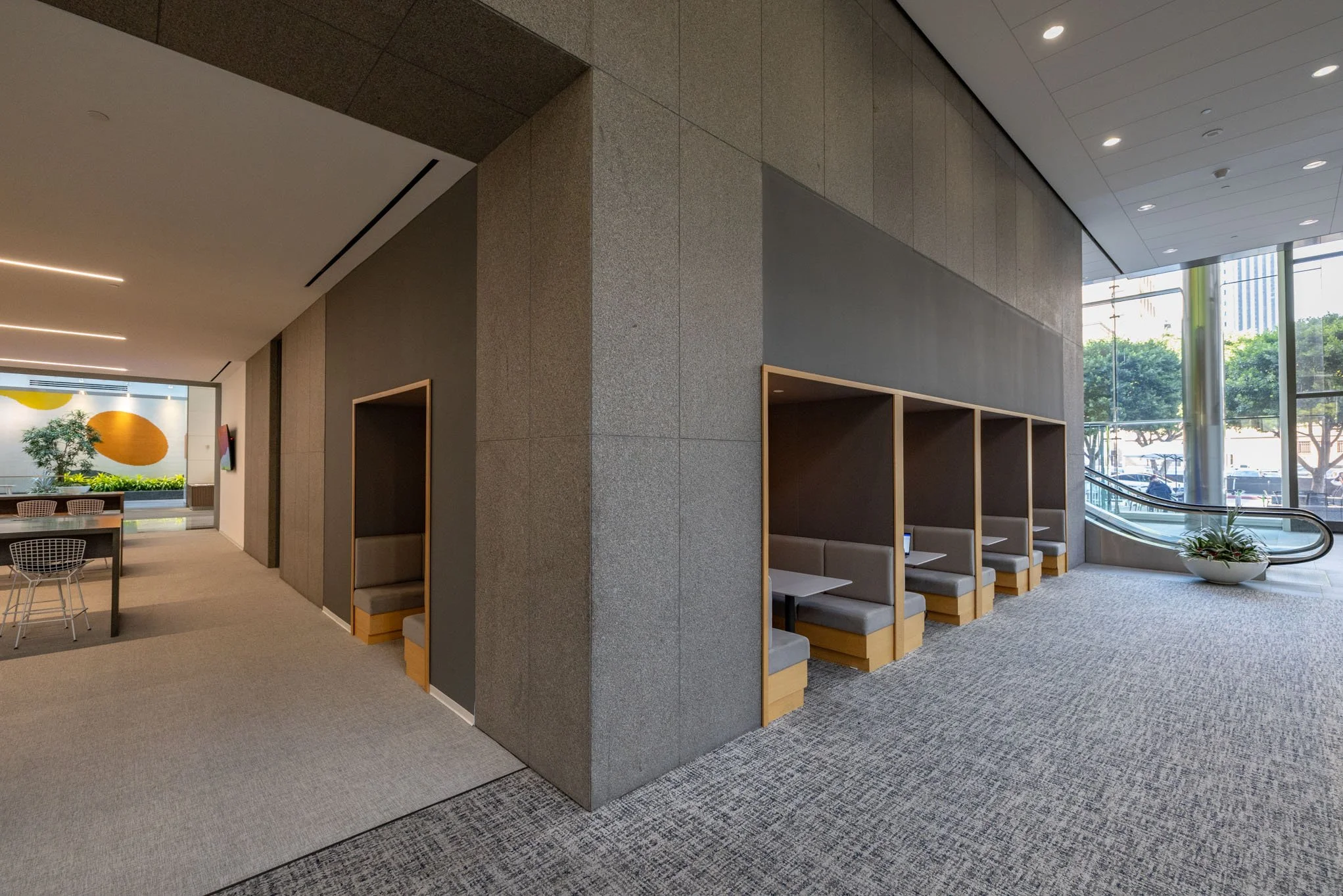 Modern indoor seating area with semi-private booths, a large window showing trees outside, and an escalator nearby.