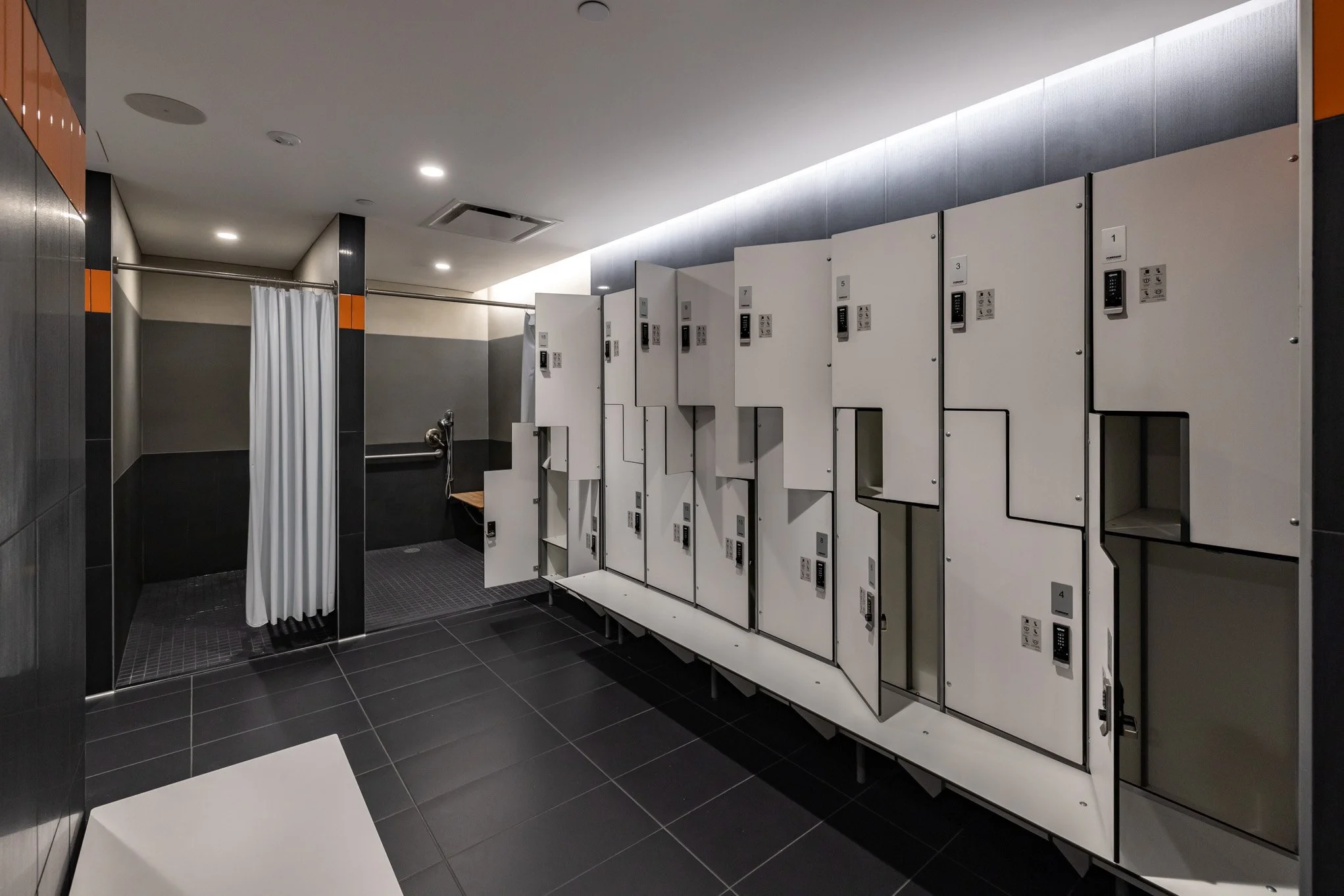 Public fitness locker room with white lockers, a changing area with a curtain, and a bench.