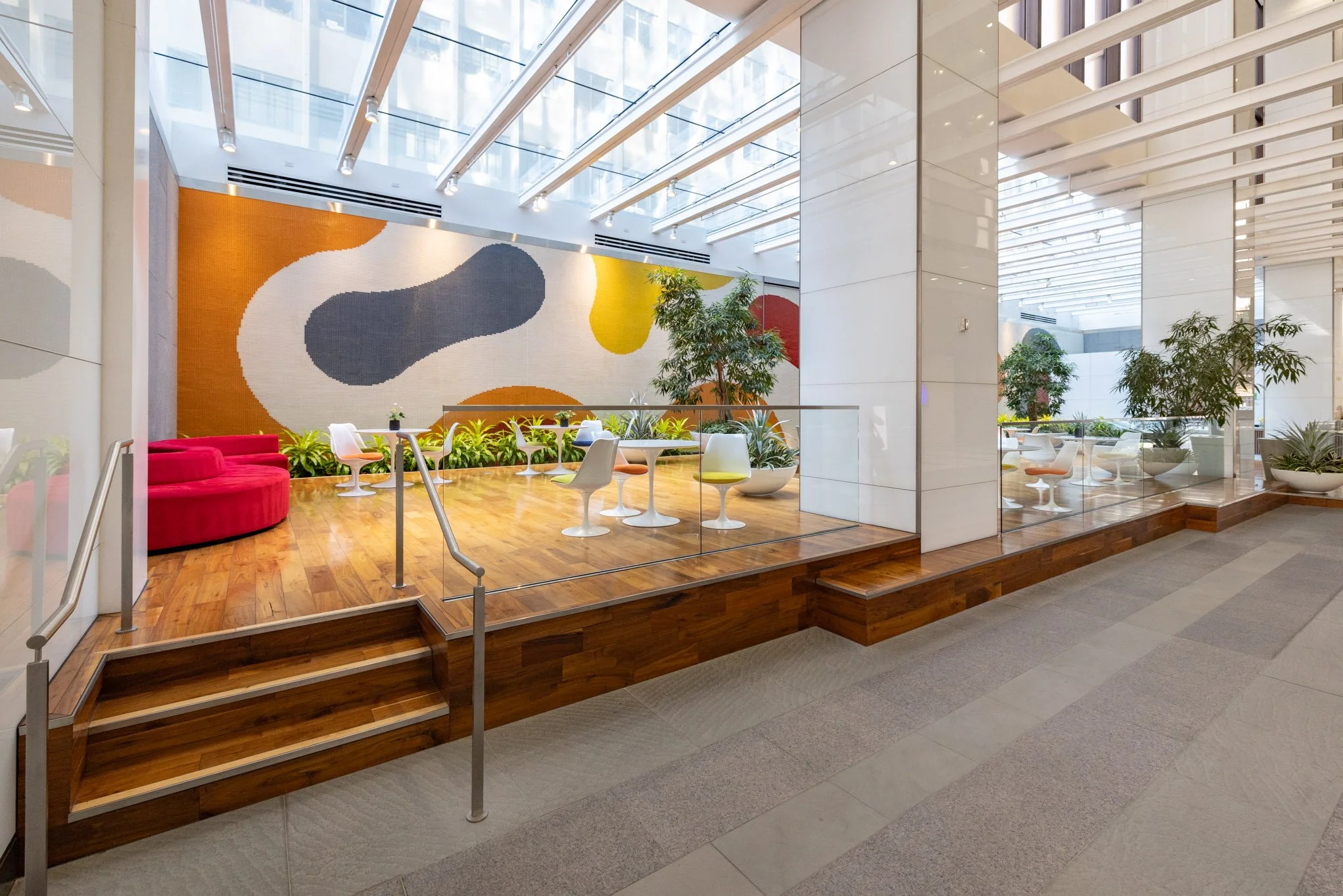 Modern indoor lounge area with wooden floor, white chairs, a red sofa, potted plants, and large colorful abstract art on the wall.