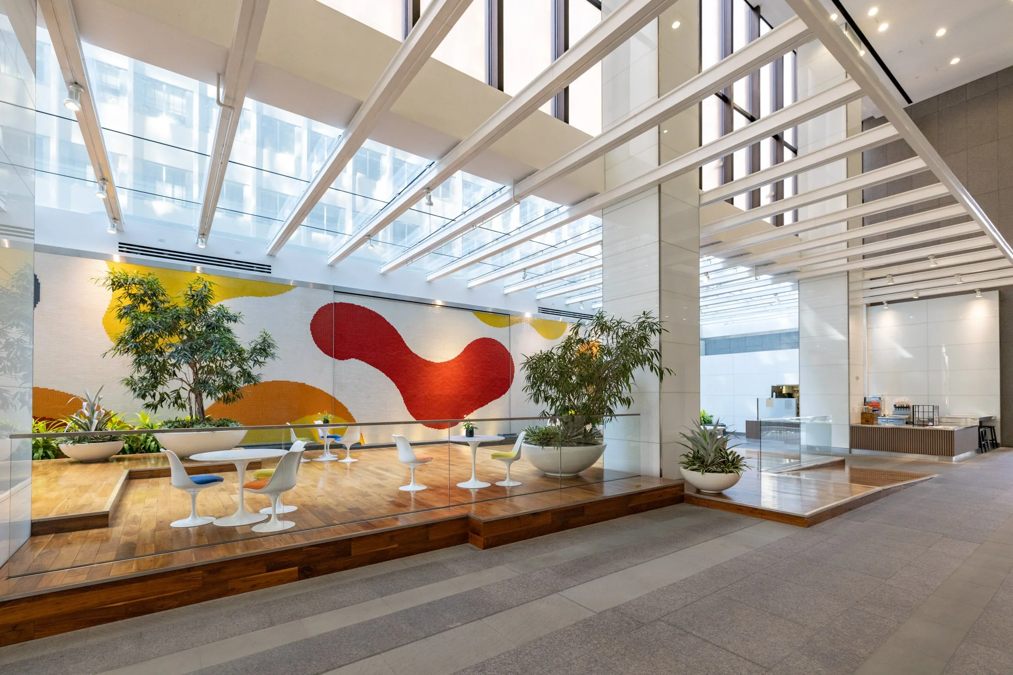 Modern indoor lounge area with round tables, white chairs, potted plants, and an abstract colorful mural on the wall.