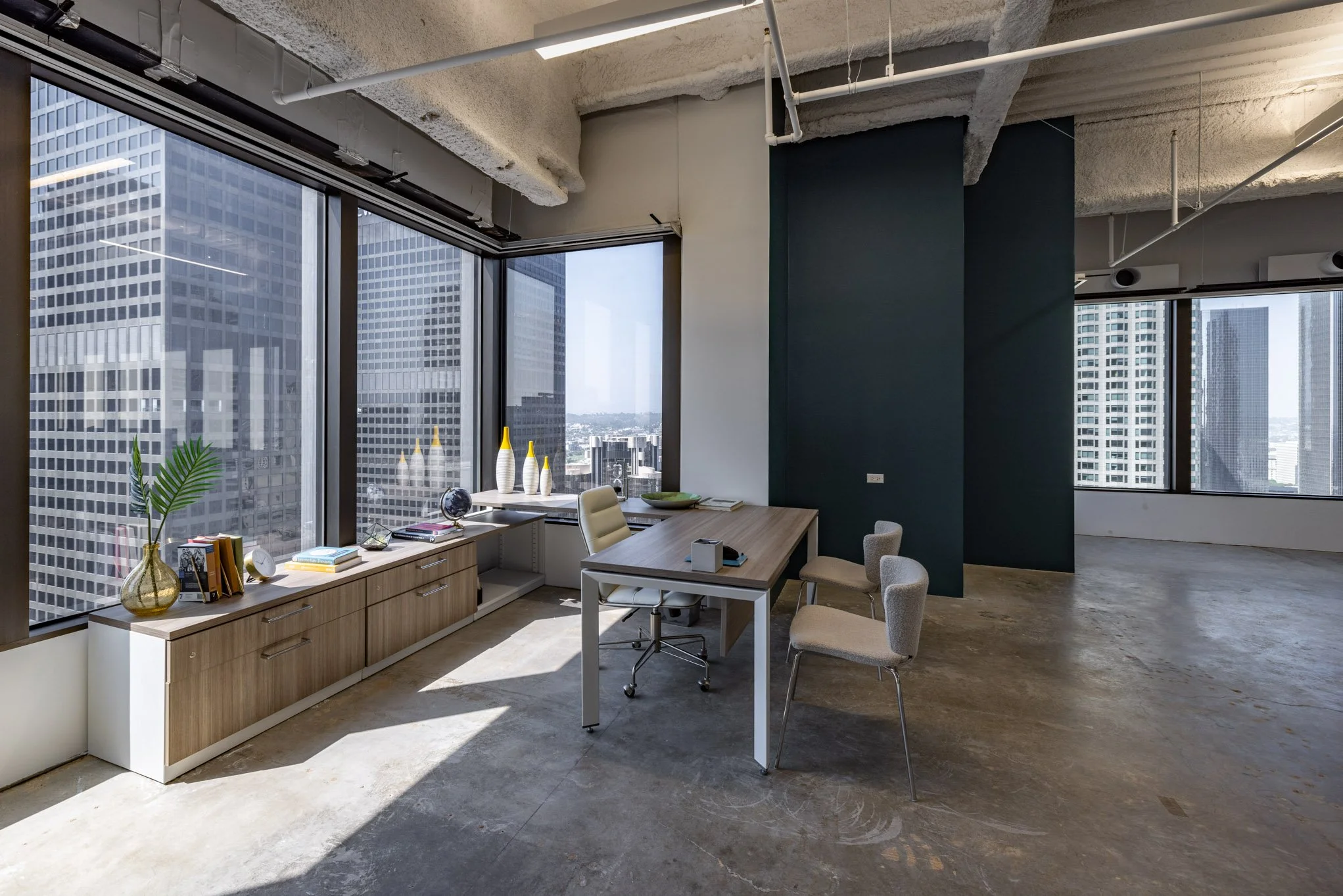 Modern office space with large windows, a wooden sideboard with decor, a small table with two chairs, and city skyscrapers outside.