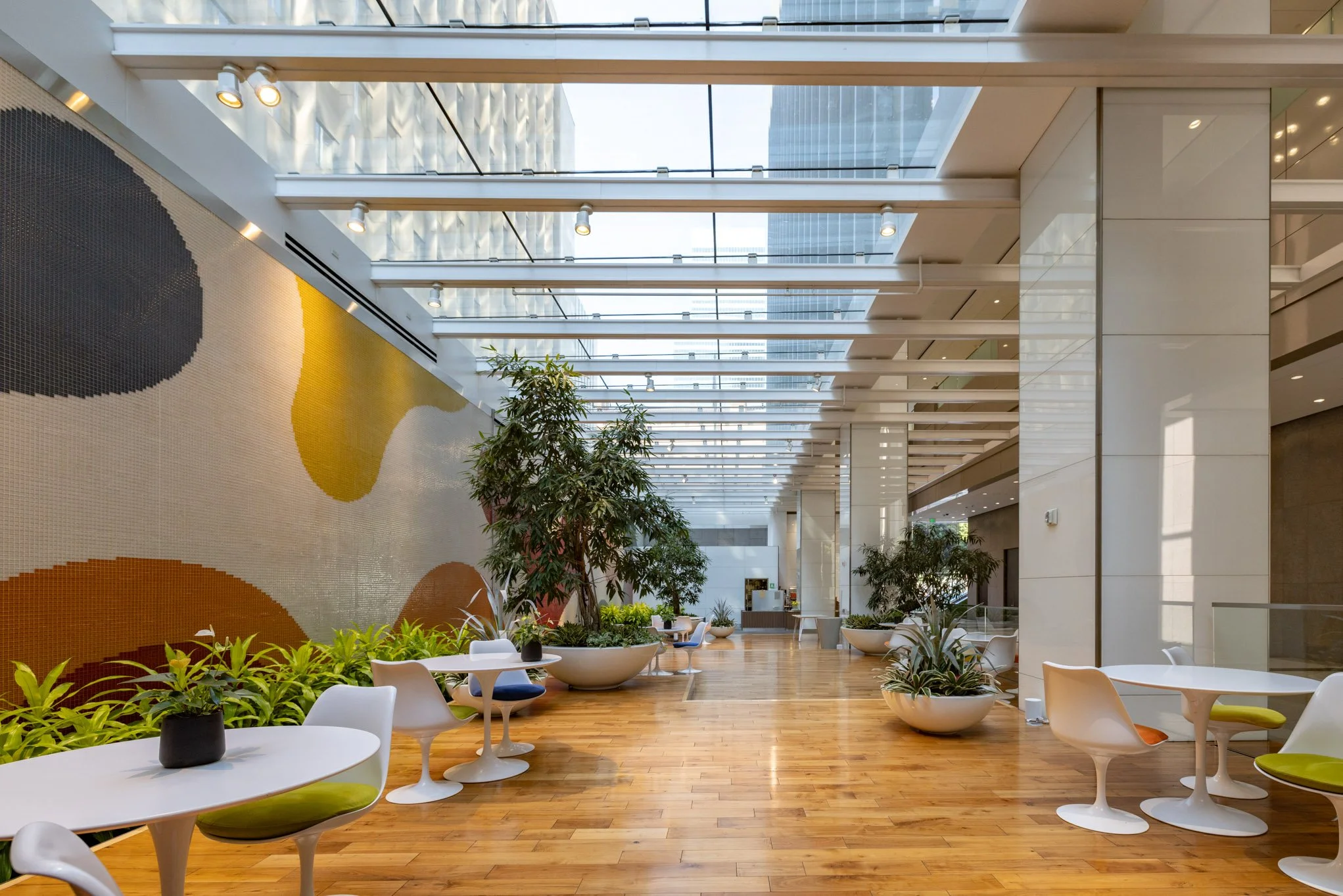Modern indoor lounge area with white tables, colorful chairs, and potted green plants, illuminated by natural light through large glass ceiling panels.