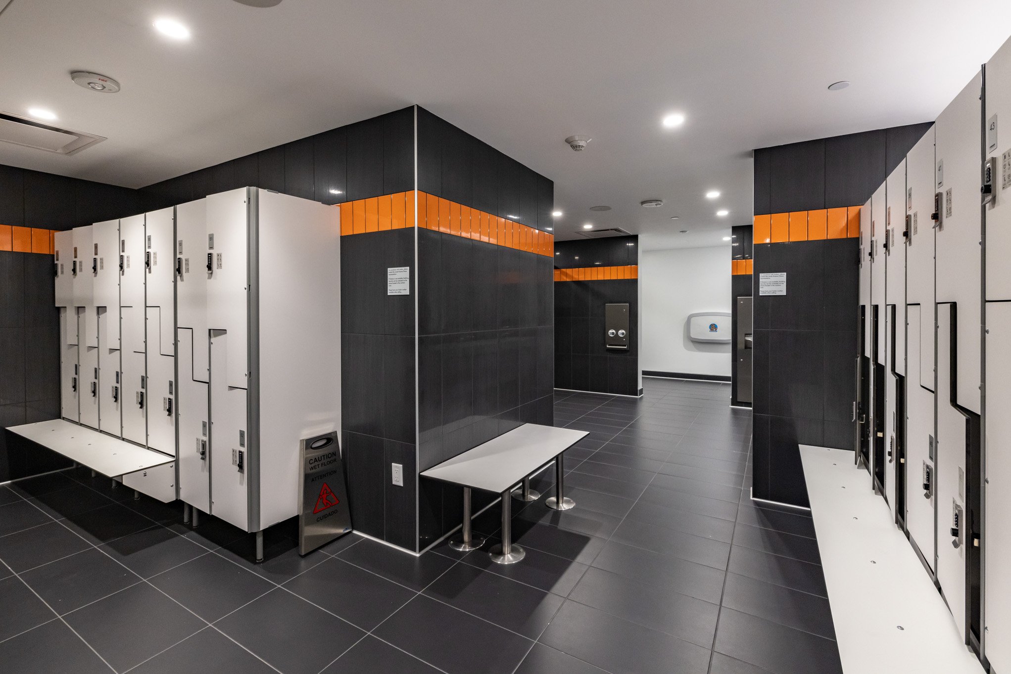 Modern locker room with white lockers, black tiled floor, dark black walls with orange accents, a wall-mounted hand sanitizer dispenser, and a small white bench.