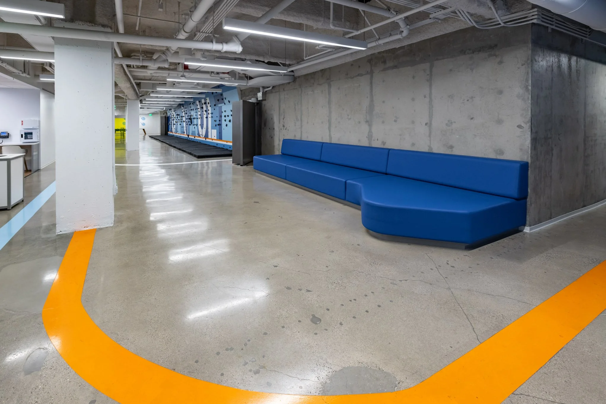 Empty indoor lobby with a long blue curved bench along a concrete wall, polished concrete floor with orange and blue directional lines, exposed pipes on ceiling, and a climbing wall in the background.