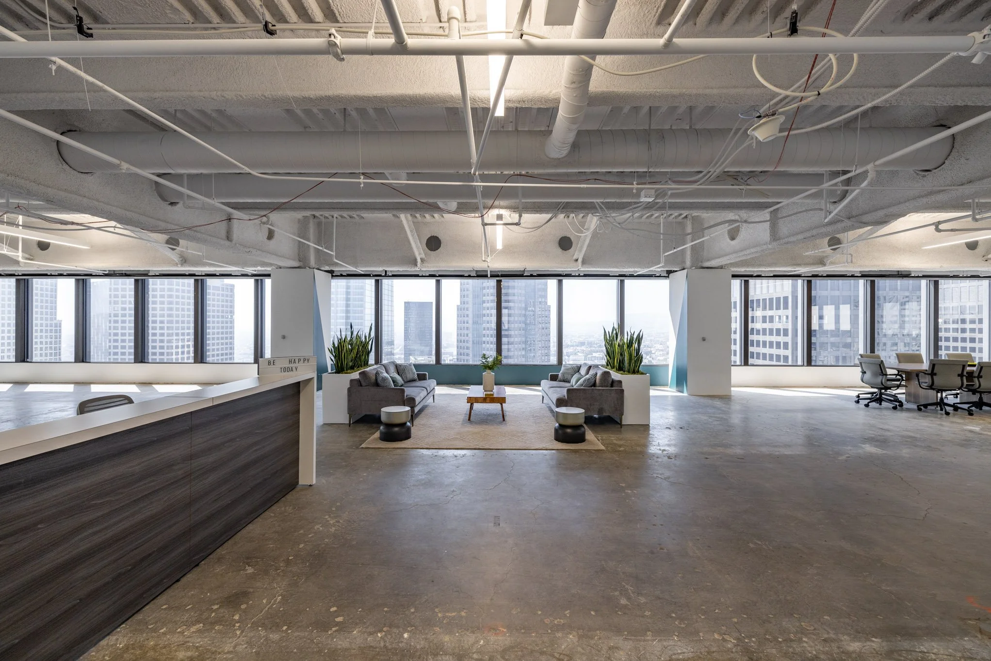 Modern office lounge area with two sofas, potted plants, a small coffee table, large windows with city view, and a spacious open floor plan.