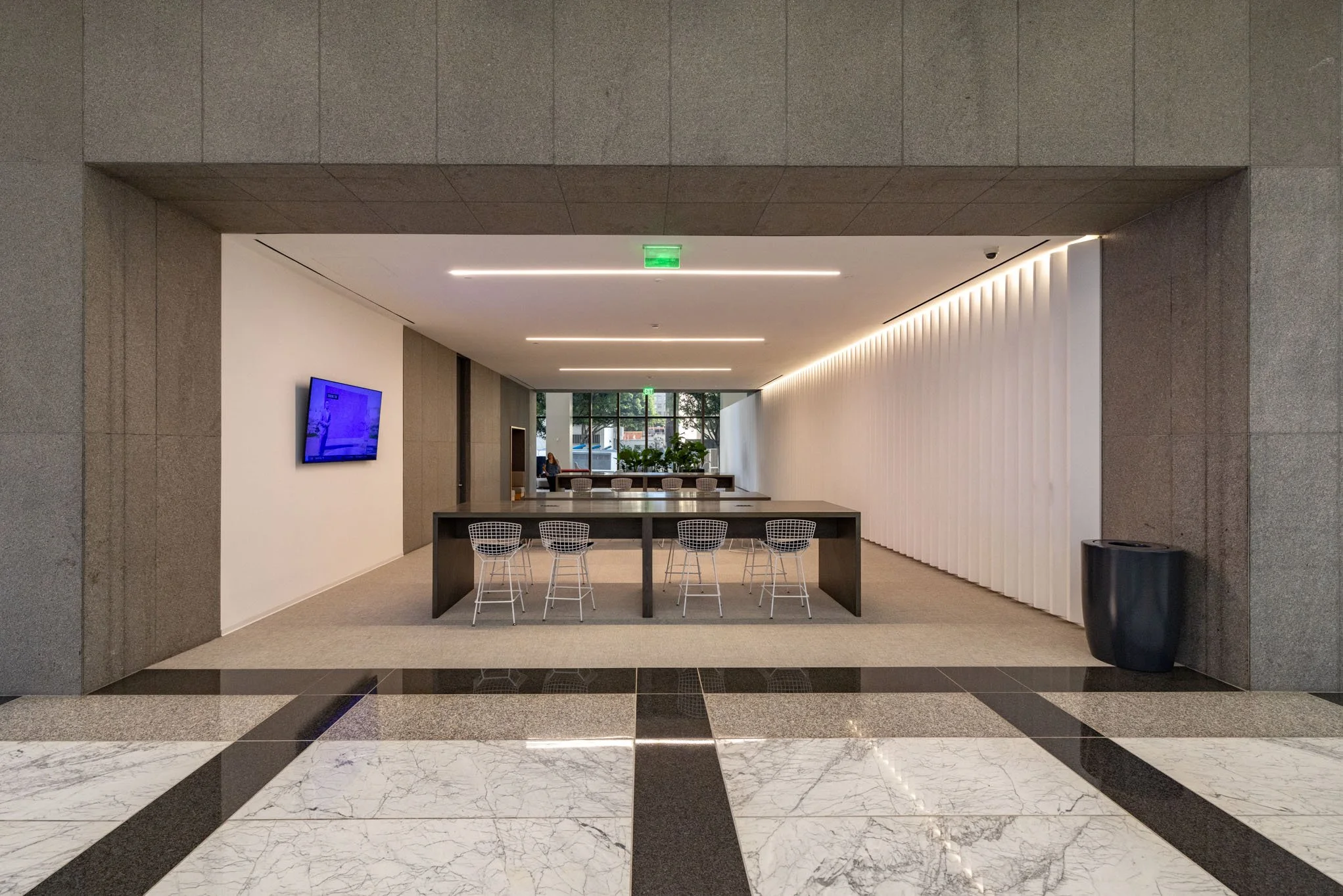 Modern office lobby with a sitting area, white and gray walls, and a large window at the back. A black table with white wire chairs is in the center, with a mounted television on the white wall to the left. Marble flooring with dark accents is in the