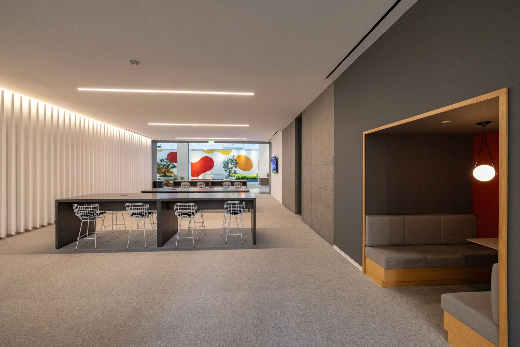 Modern interior space with a long high table and wire chairs, a booth with seating, and a window decorated with colorful shapes.
