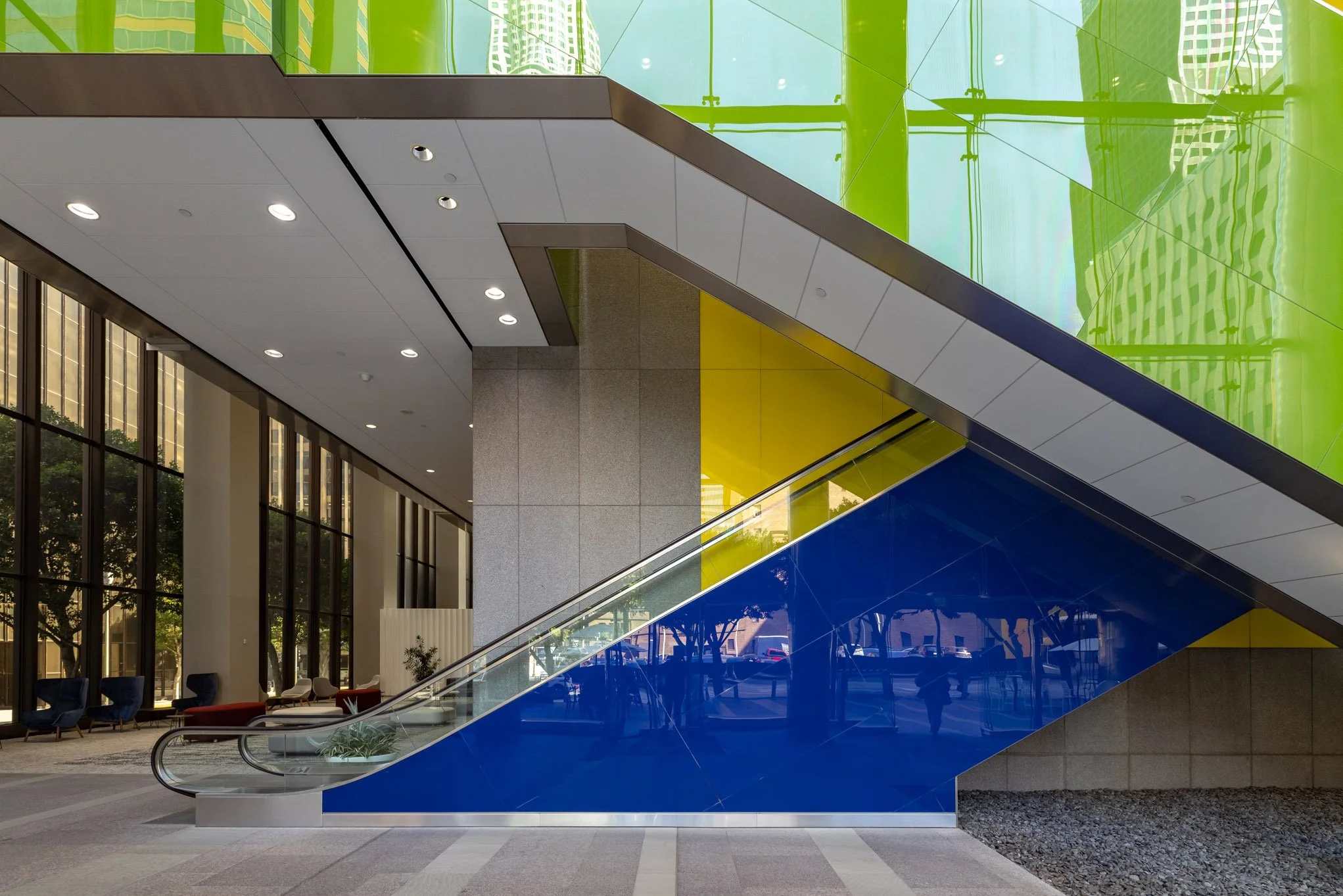 Inside a modern building with an escalator painted blue and yellow, large glass windows, and seating areas.
