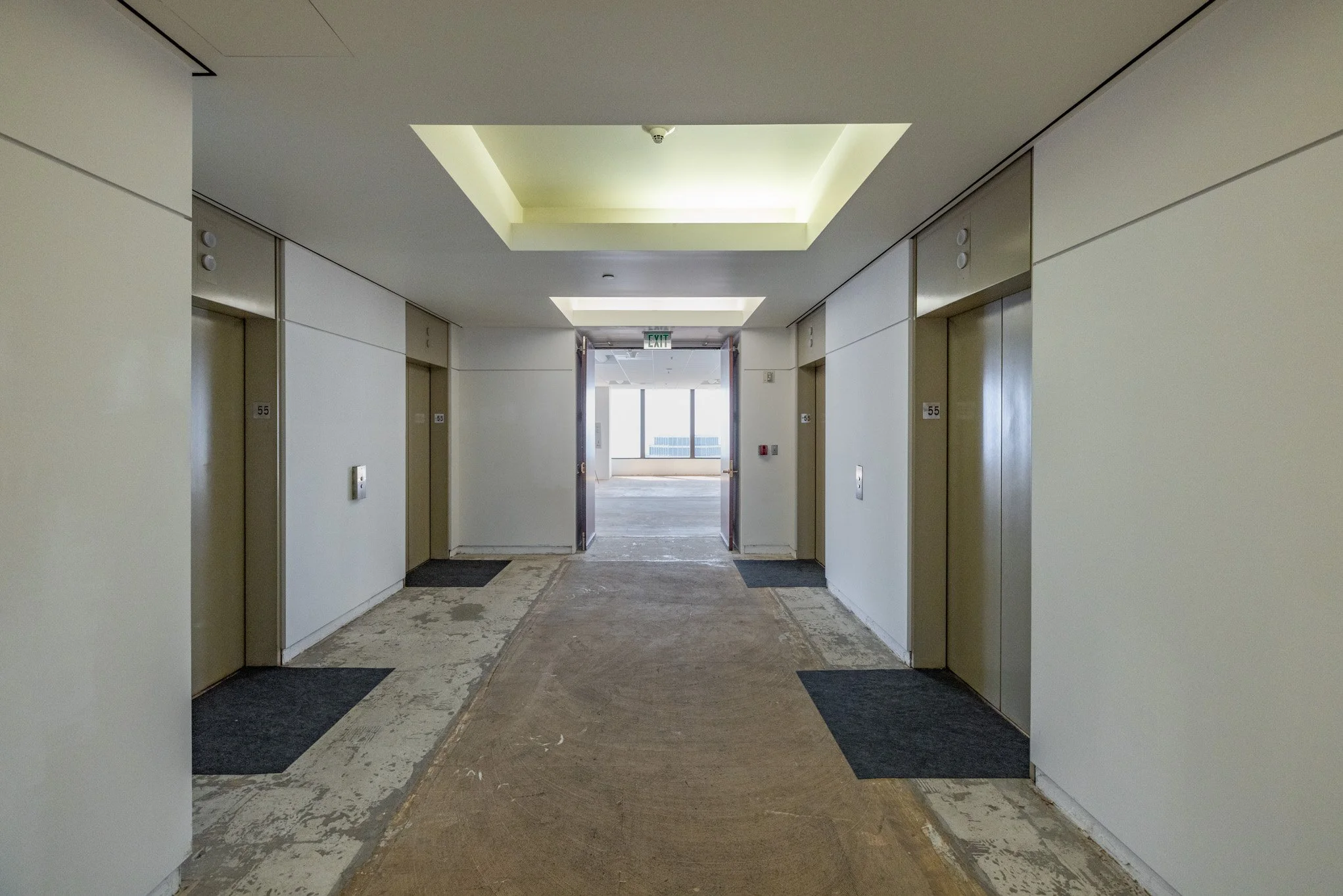 Empty hallway with elevators on either side, carpeted and under renovation, leading to a bright area with windows at the end.