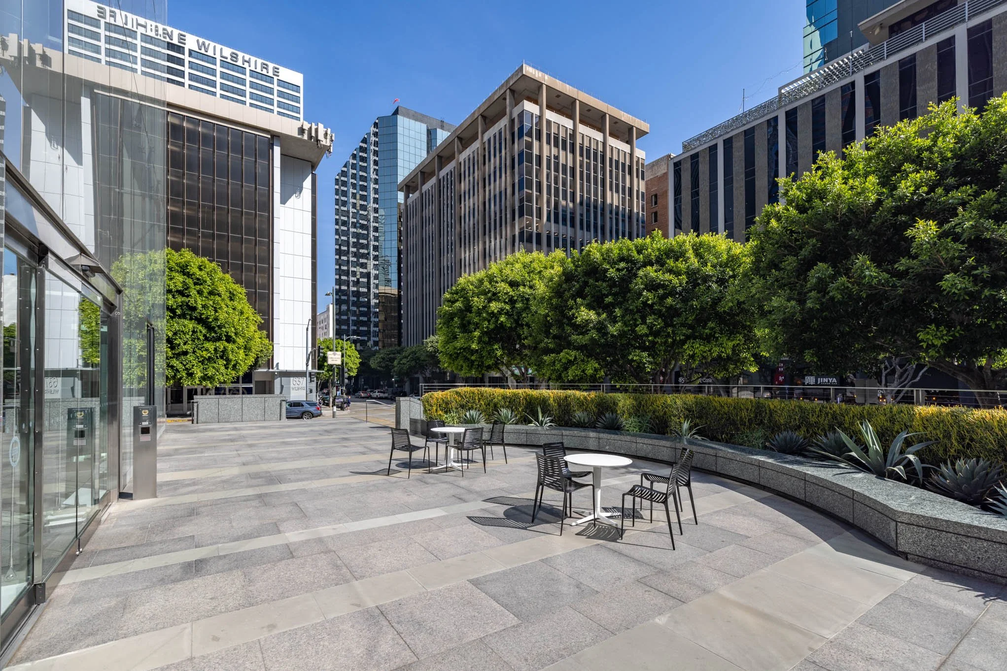 An outdoor area with a few tables and chairs, surrounded by greenery, in an urban cityscape with tall glass office buildings under a clear blue sky.