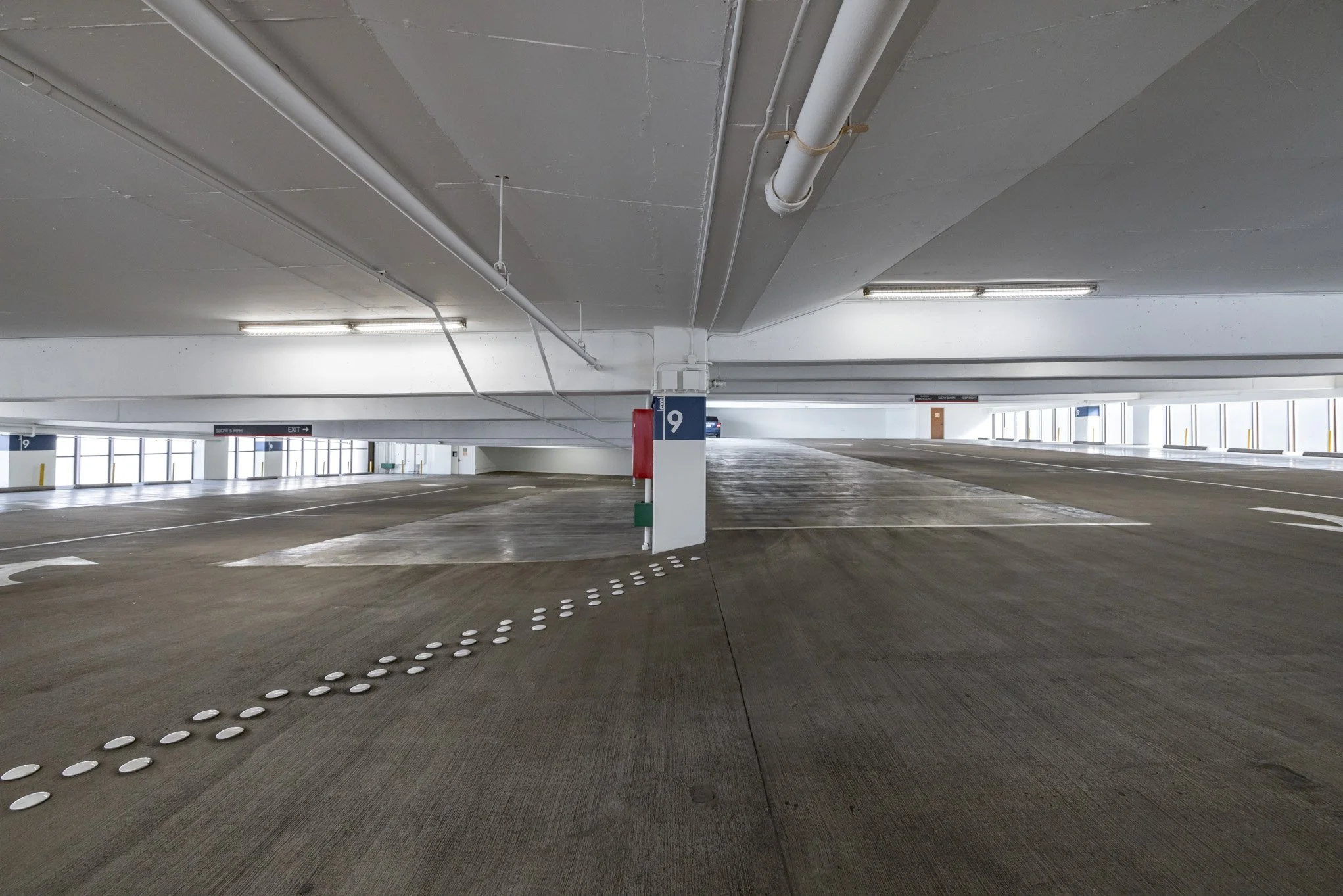 Empty multi-level parking garage with concrete floors, white walls, and ceiling pipes, illuminated by fluorescent lights.