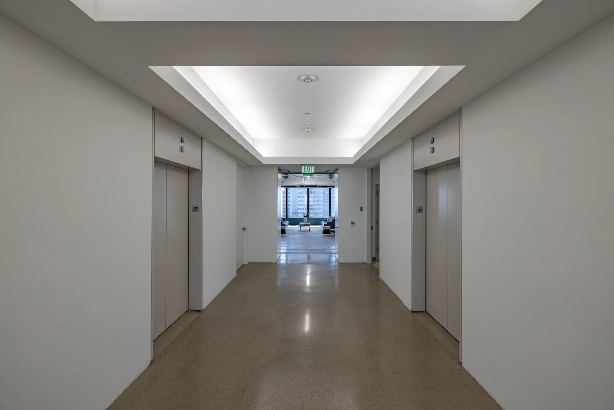 Hallway with two elevator doors on each side, leading to a lounge area with large windows and city view, at the end of the hallway.