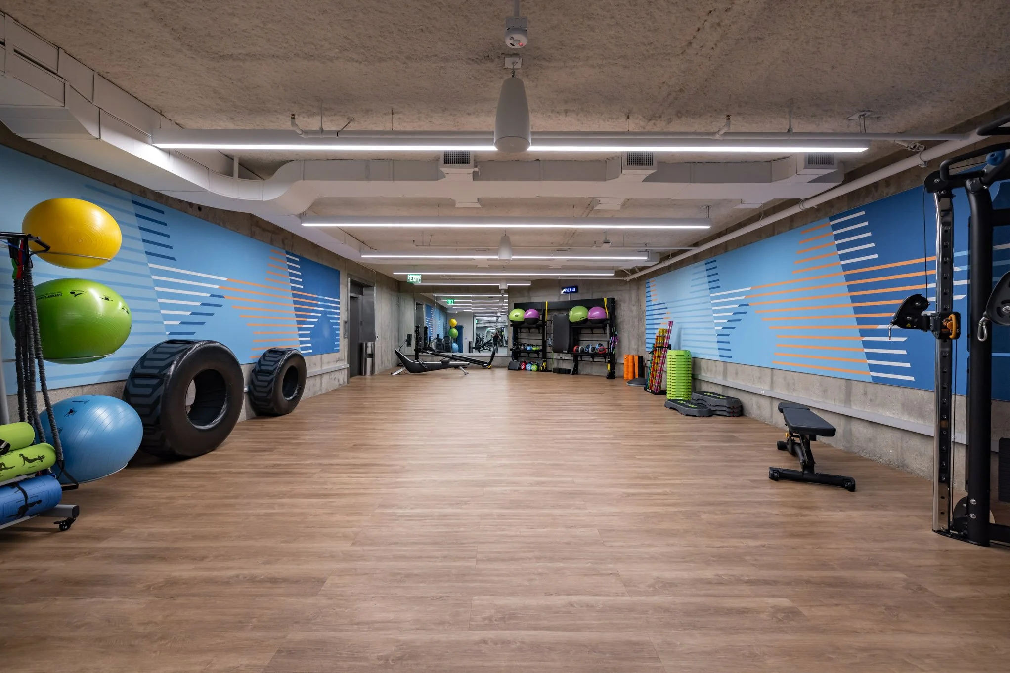 Empty fitness gym with wooden floors, blue wall murals, and various workout equipment including stability balls, tires, weights, and exercise mats.