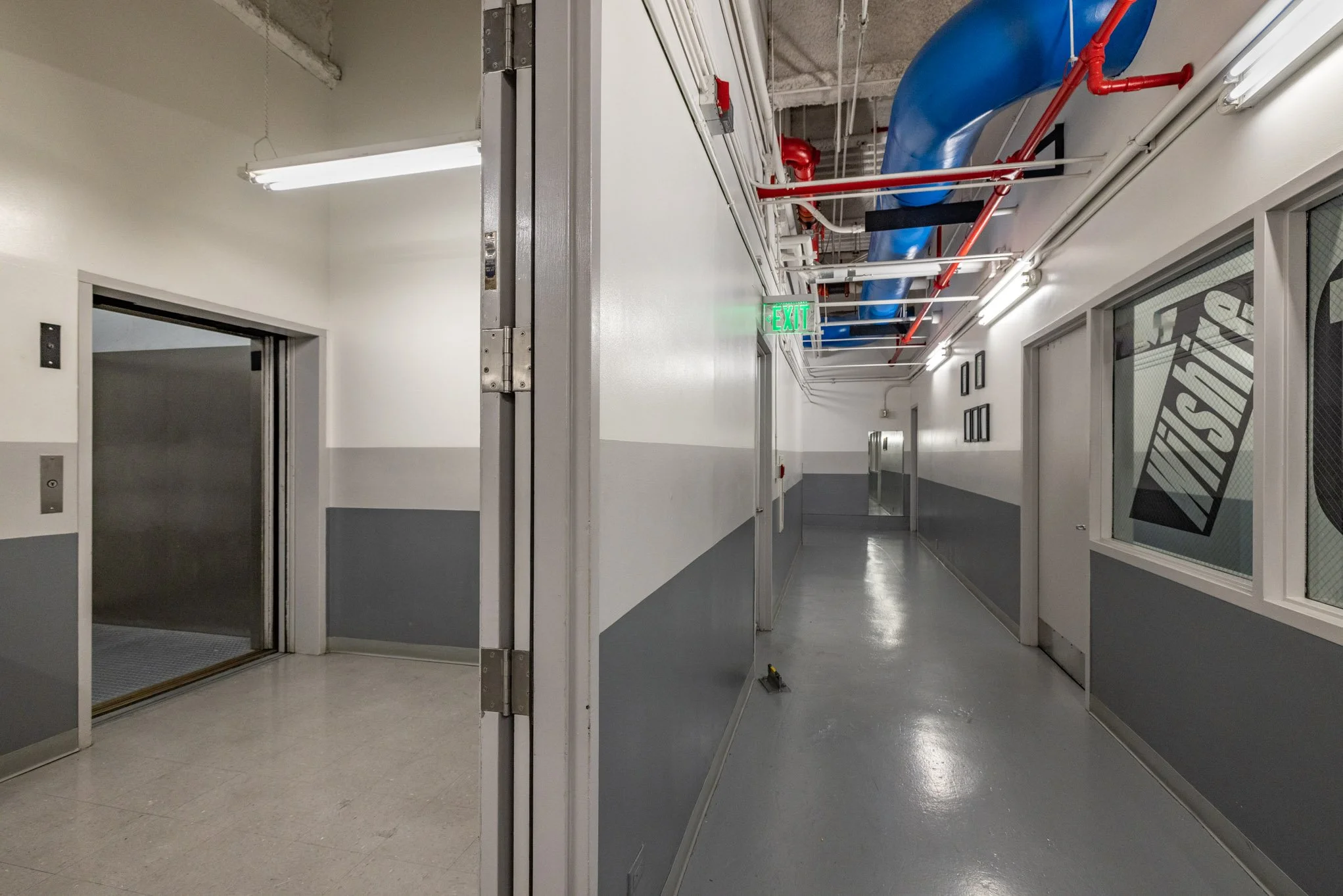 An indoor hallway with an elevator on the left and a corridor with ceiling pipes and ventilation ducts, illuminated by fluorescent lights.