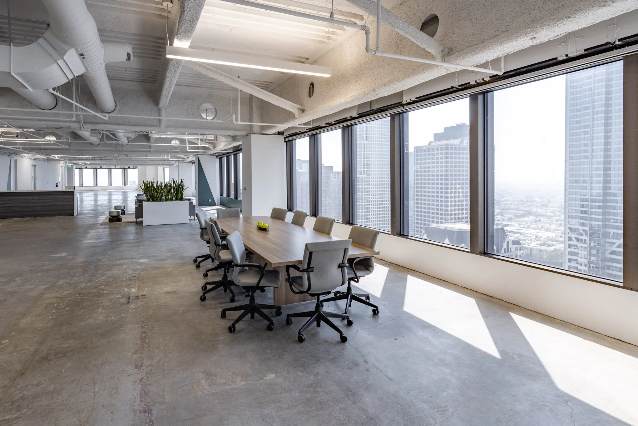 Modern office conference room with large windows, a long wooden table, and gray swivel chairs, overlooking a city skyline.