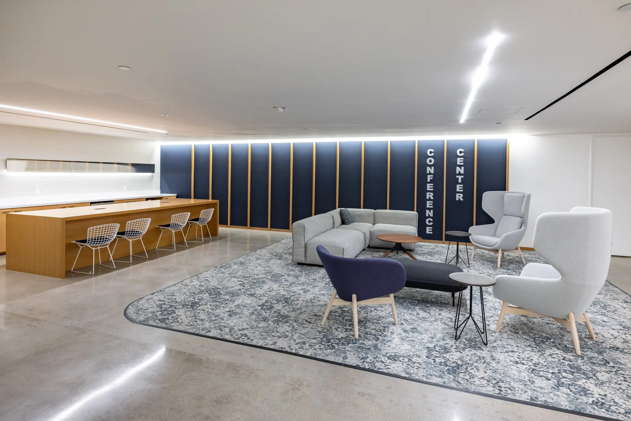 Modern conference center lounge with a seating area including a gray sectional sofa, various armchairs, and small tables, with a dark blue accent wall labeled 'Conference Center' in the background and a wooden counter with wire chairs on the left.