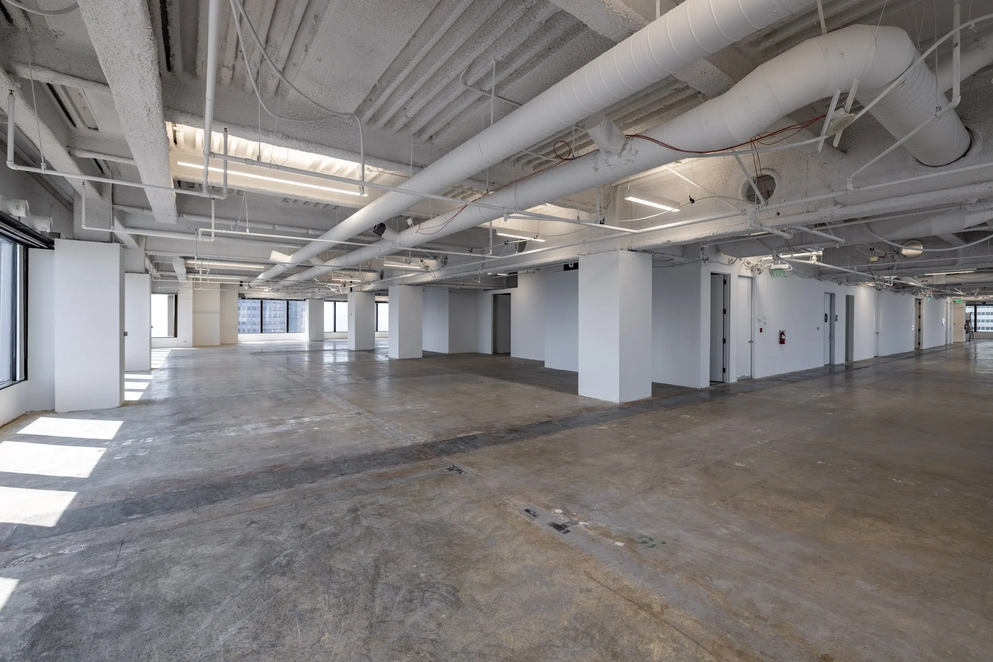 Empty commercial office space with unfinished concrete floor, white walls, large windows, and exposed ceiling with white ducts and pipes.